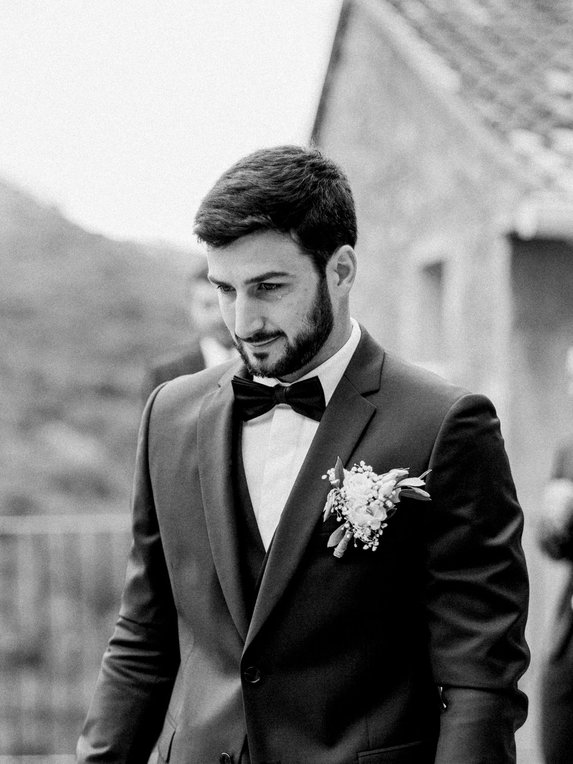 Black and white photo of a groom in a suit with bow tie and boutonniere, looking down thoughtfully.