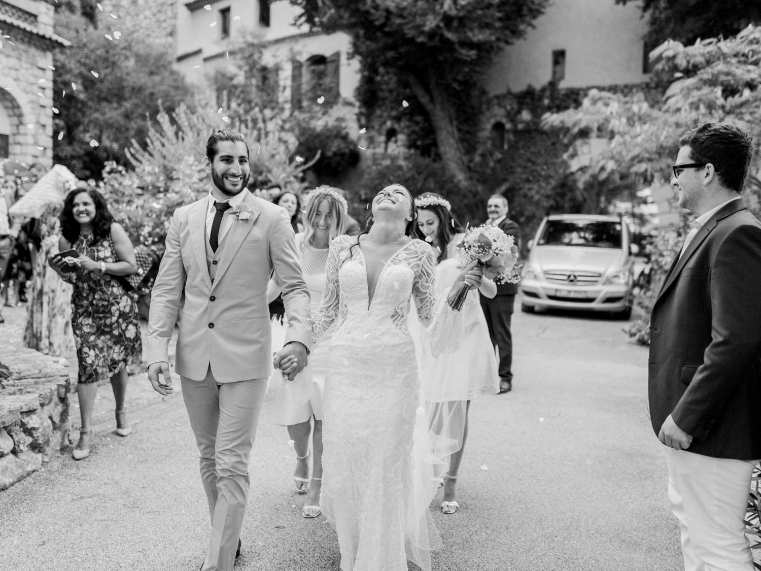 Black and white photo of a smiling bride and groom walking hand in hand with wedding guests celebrating outdoors.