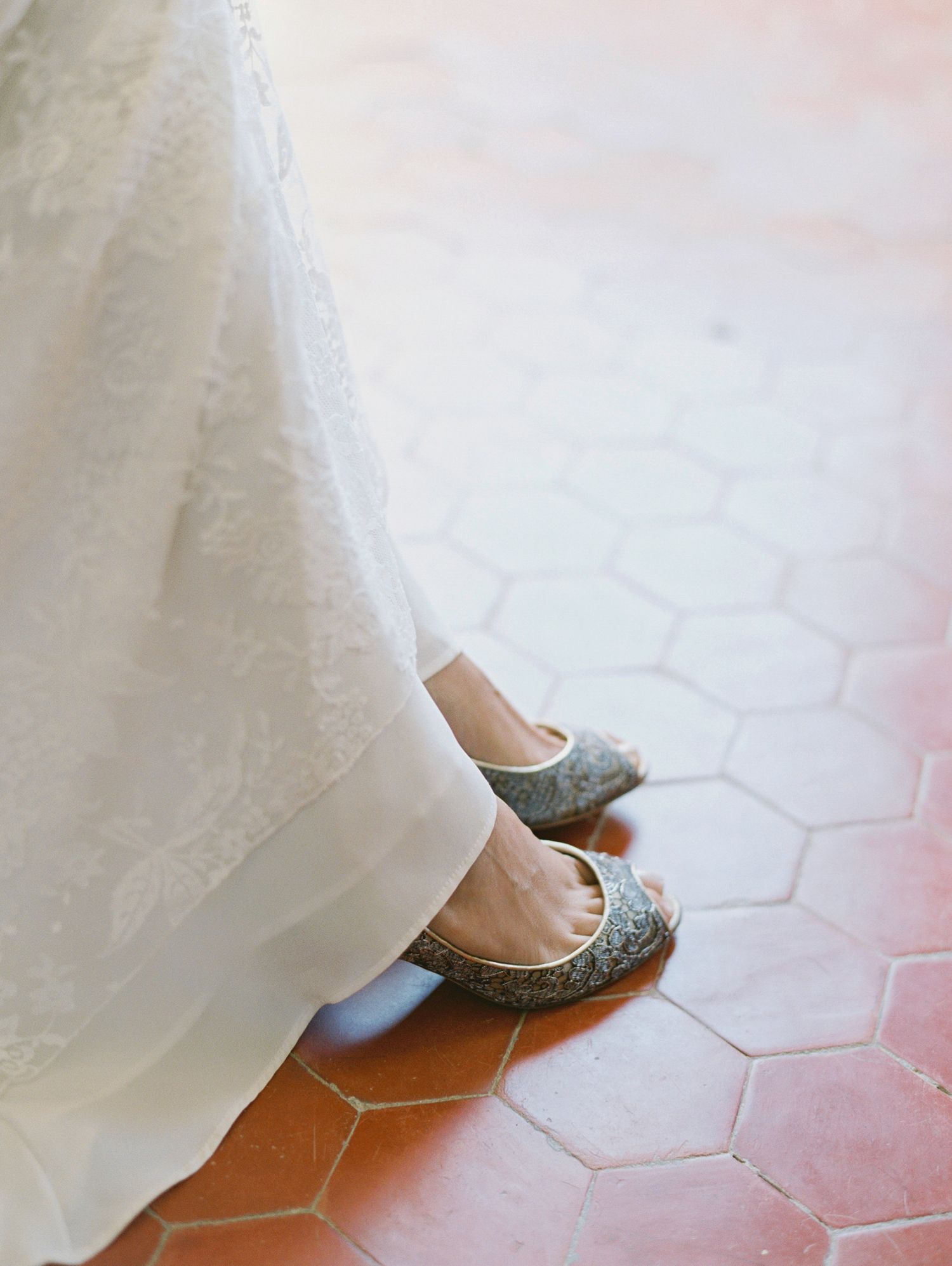 Bride wearing glittery gold flats under a white lace wedding gown on terracotta hexagon tile floor.