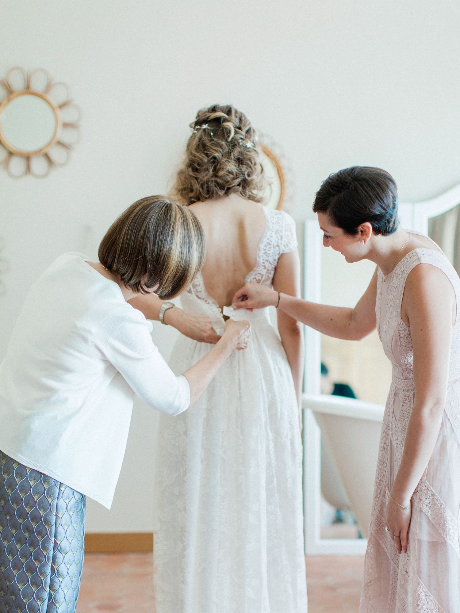 Two women helping a bride button her lace wedding dress from behind near a mirror.