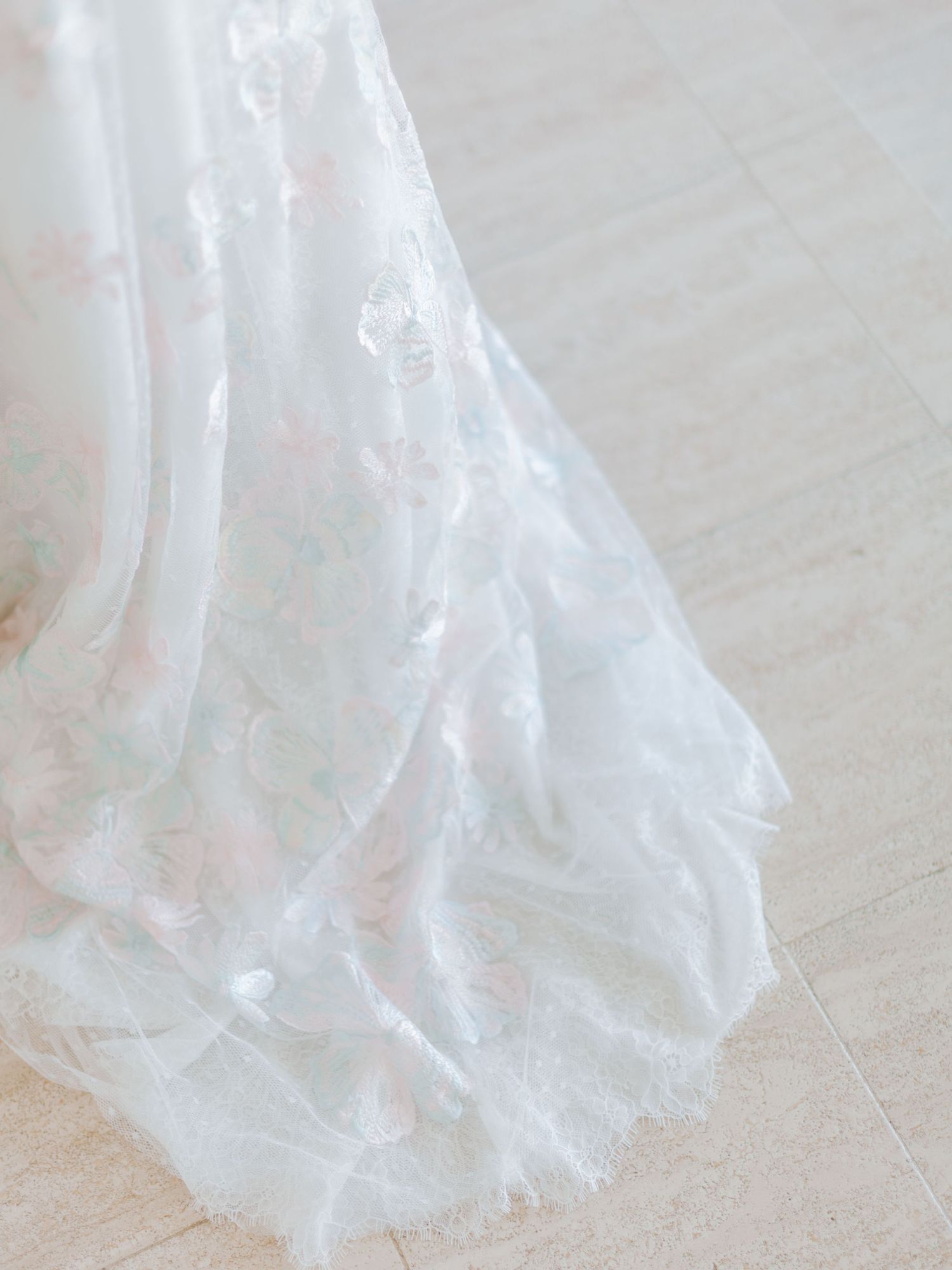 Traîne de robe de mariée en dentelle blanche délicate, ornée de doux motifs floraux et drapée avec élégance sur un sol en carrelage clair.