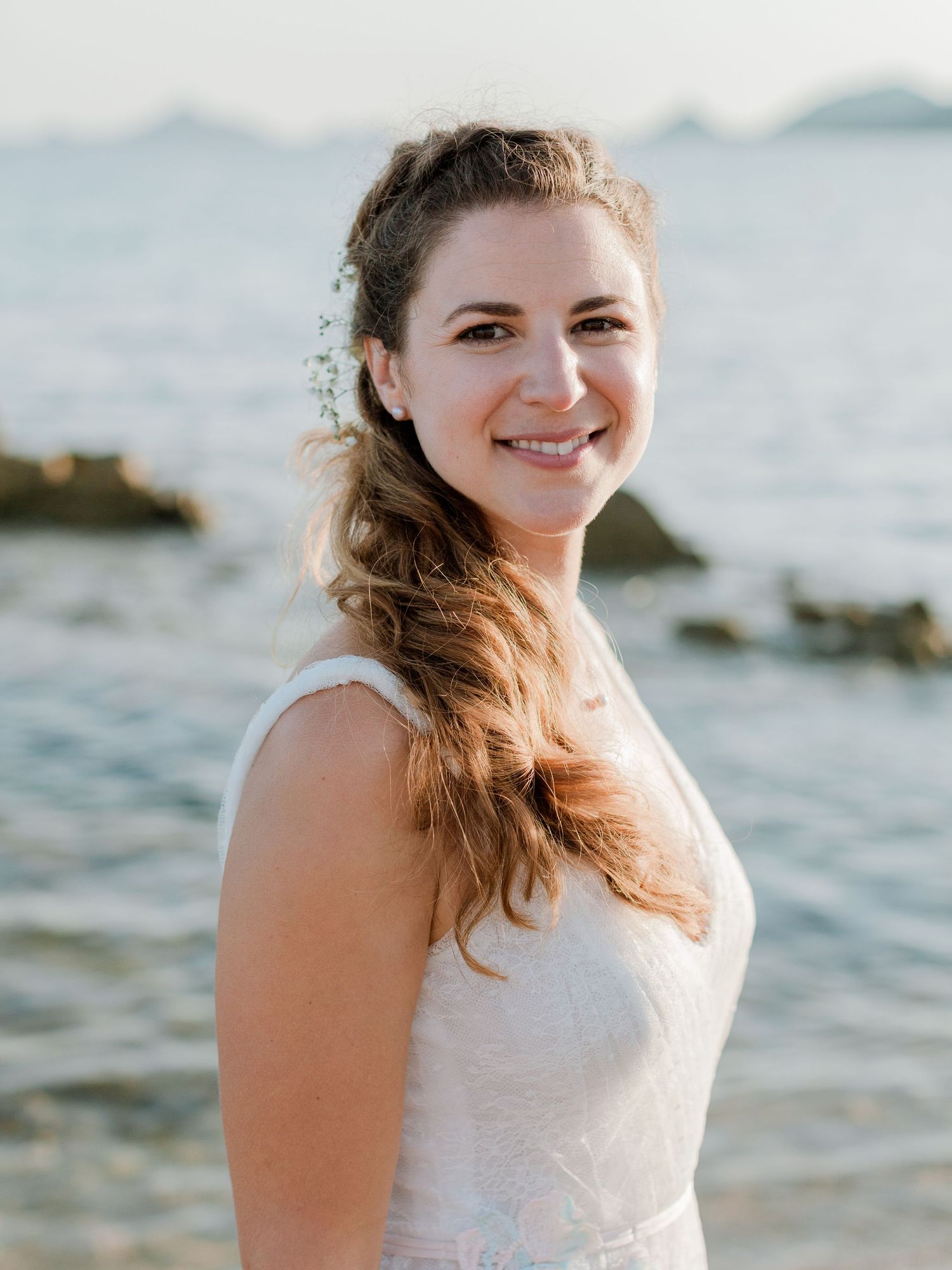 Une mariée souriante en robe blanche, portant un accessoire floral dans les cheveux, pose sur une plage rocheuse au coucher du soleil.