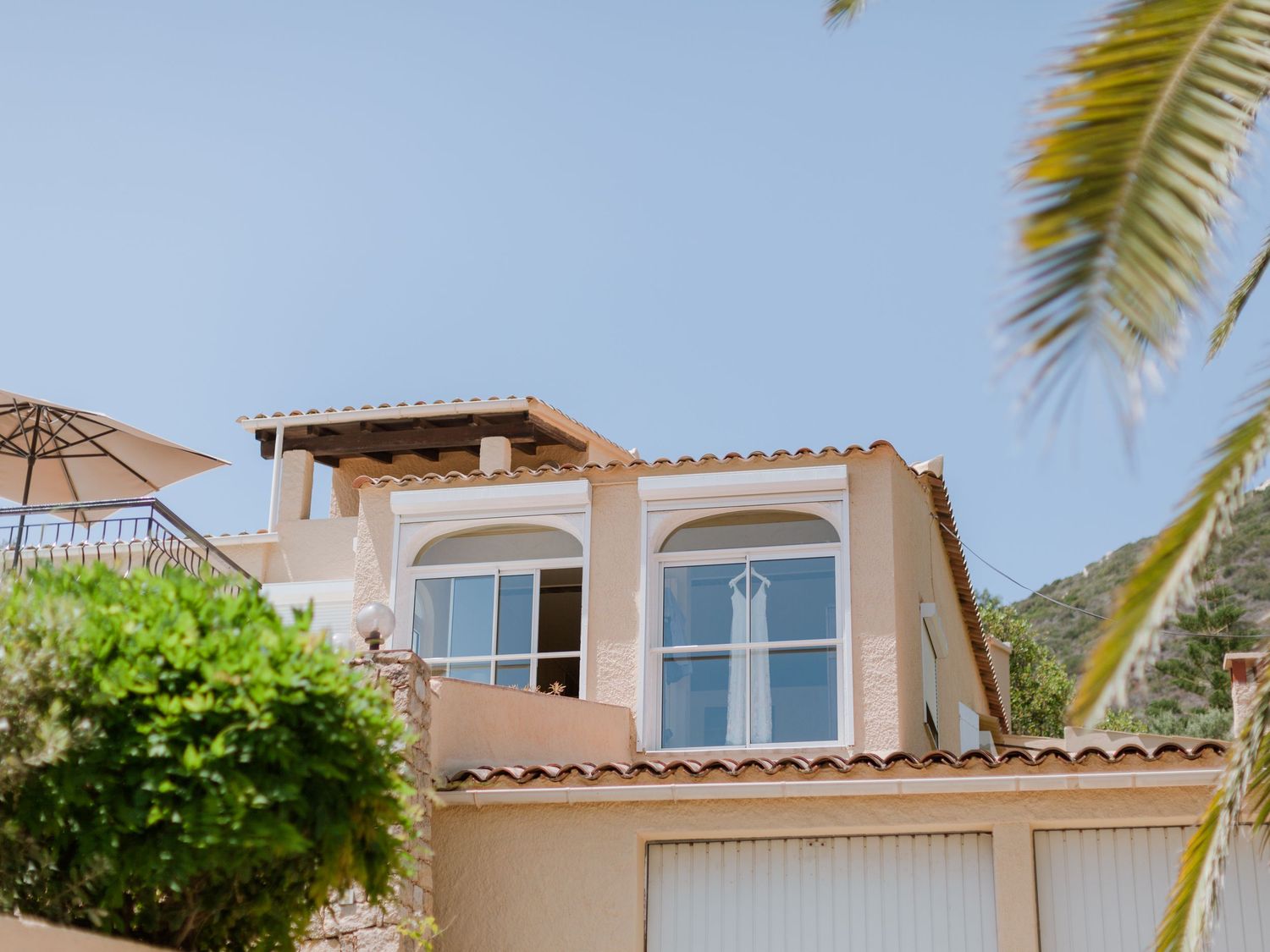 Villa de style méditerranéen avec toit en terre cuite, fenêtres en arc et palmiers se découpant sur un ciel bleu limpide.