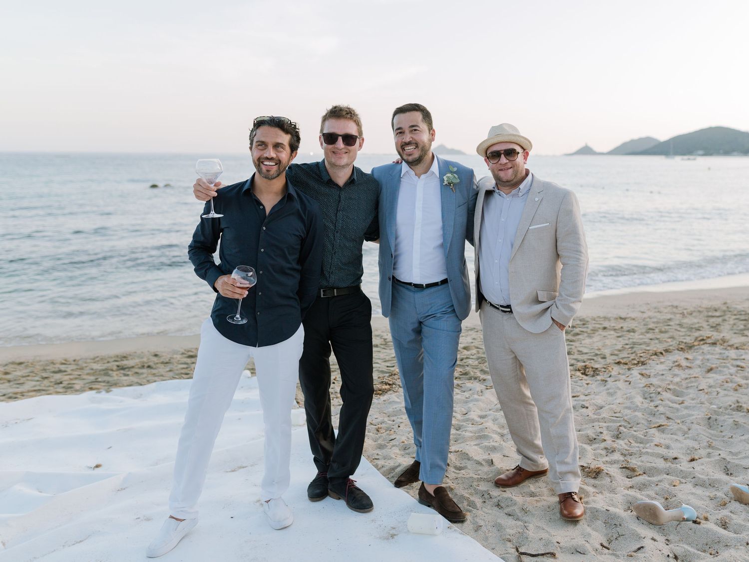 Quatre hommes posant ensemble sur une plage de sable au coucher du soleil, vêtus d'une tenue élégante mais décontractée lors d'un mariage