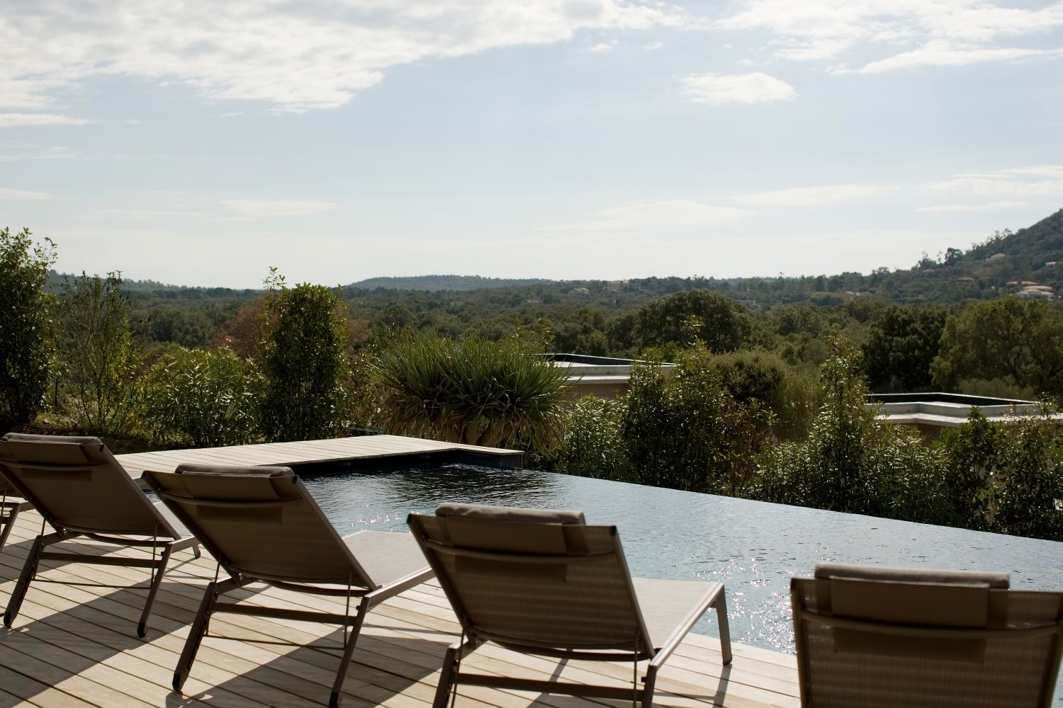 Piscine à débordement avec terrasse en bois, chaises longues donnant sur un paysage vallonné boisé sous un ciel bleu.