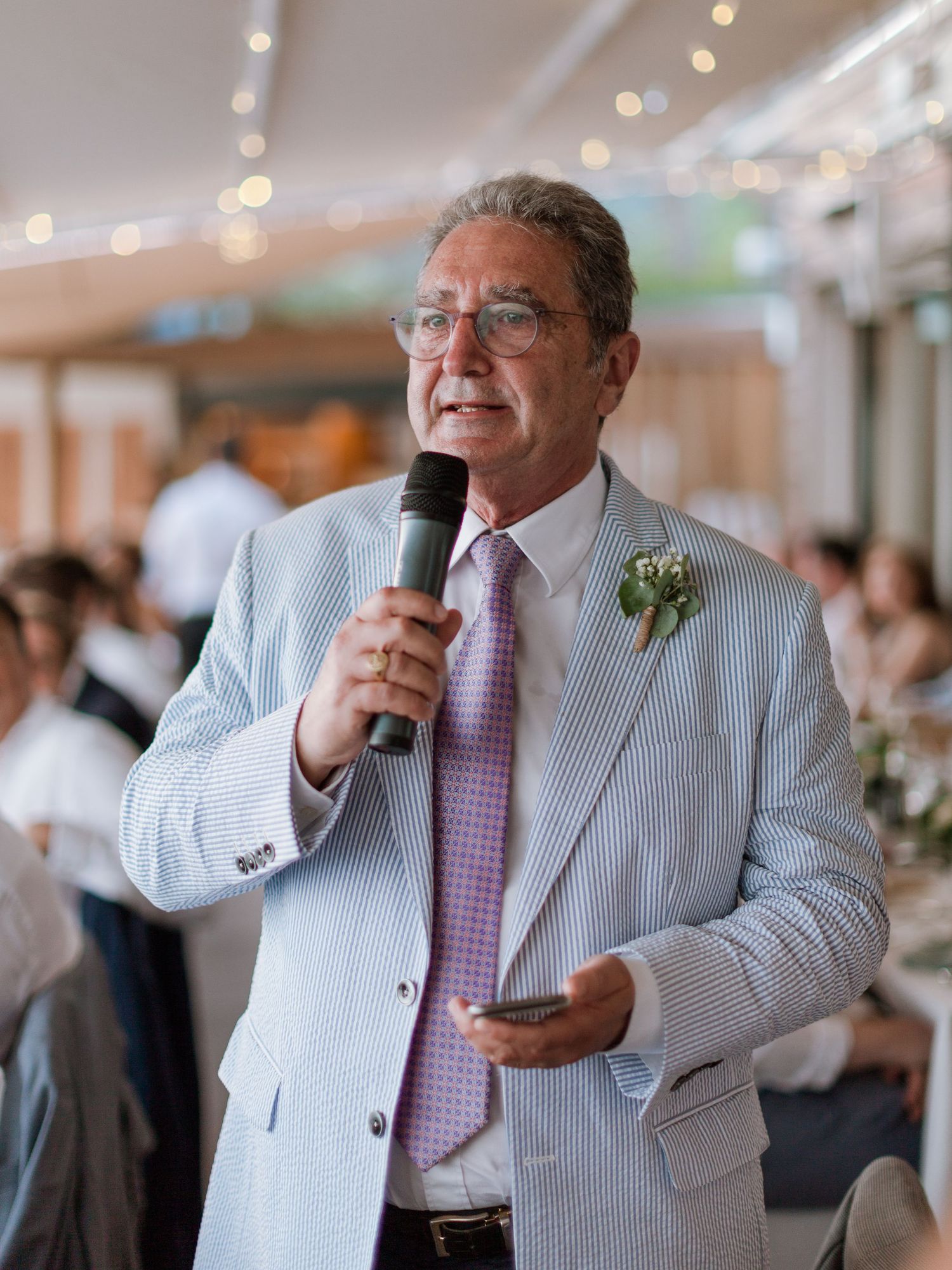 Un homme en costume en seersucker, portant une fleur à la boutonnière, prononce un discours lors d'une réception de mariage