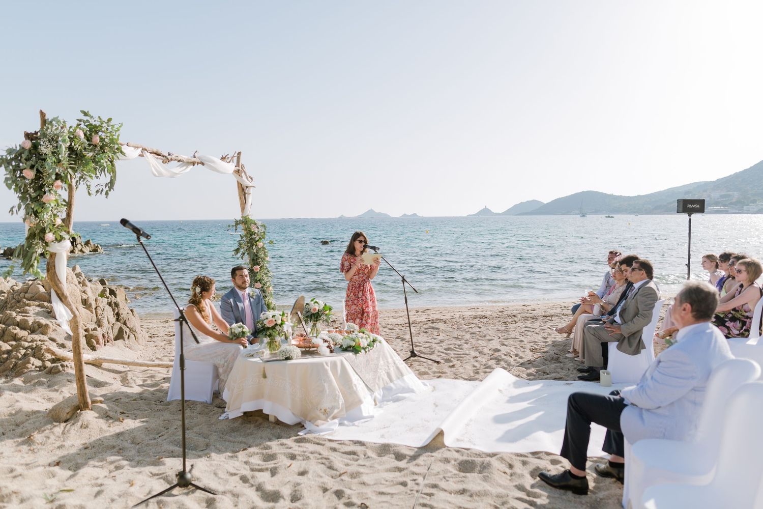 Cérémonie de mariage romantique sur la plage, avec le couple sous une arche fleurie, l'officiant et les invités assis au bord de l'océan.