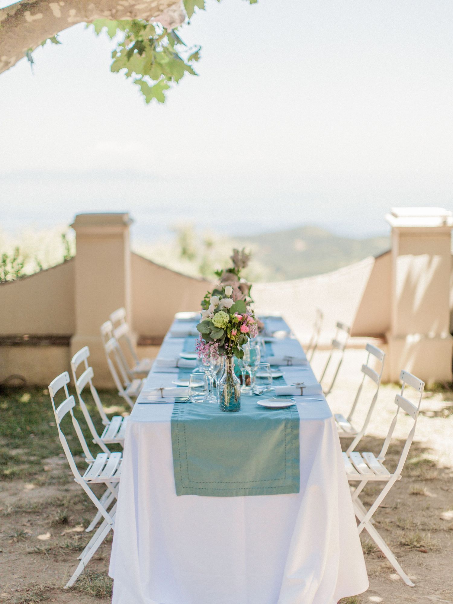 Elegant outdoor wedding reception table with blue runner, floral centerpiece, white chairs, and scenic hilltop views.