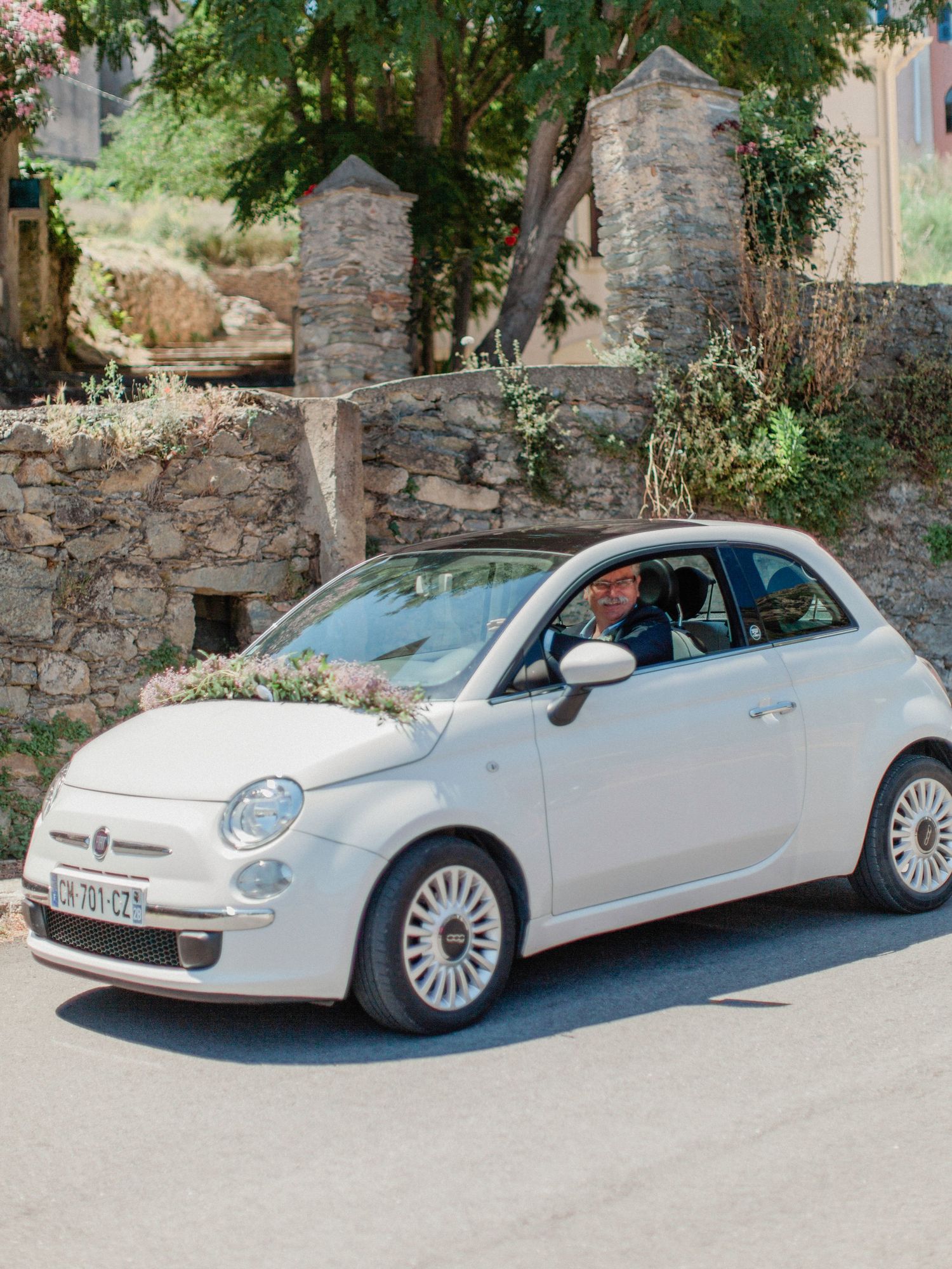 A white Fiat 500 decorated with flowers drives through a scenic stone village road on a sunny wedding day.