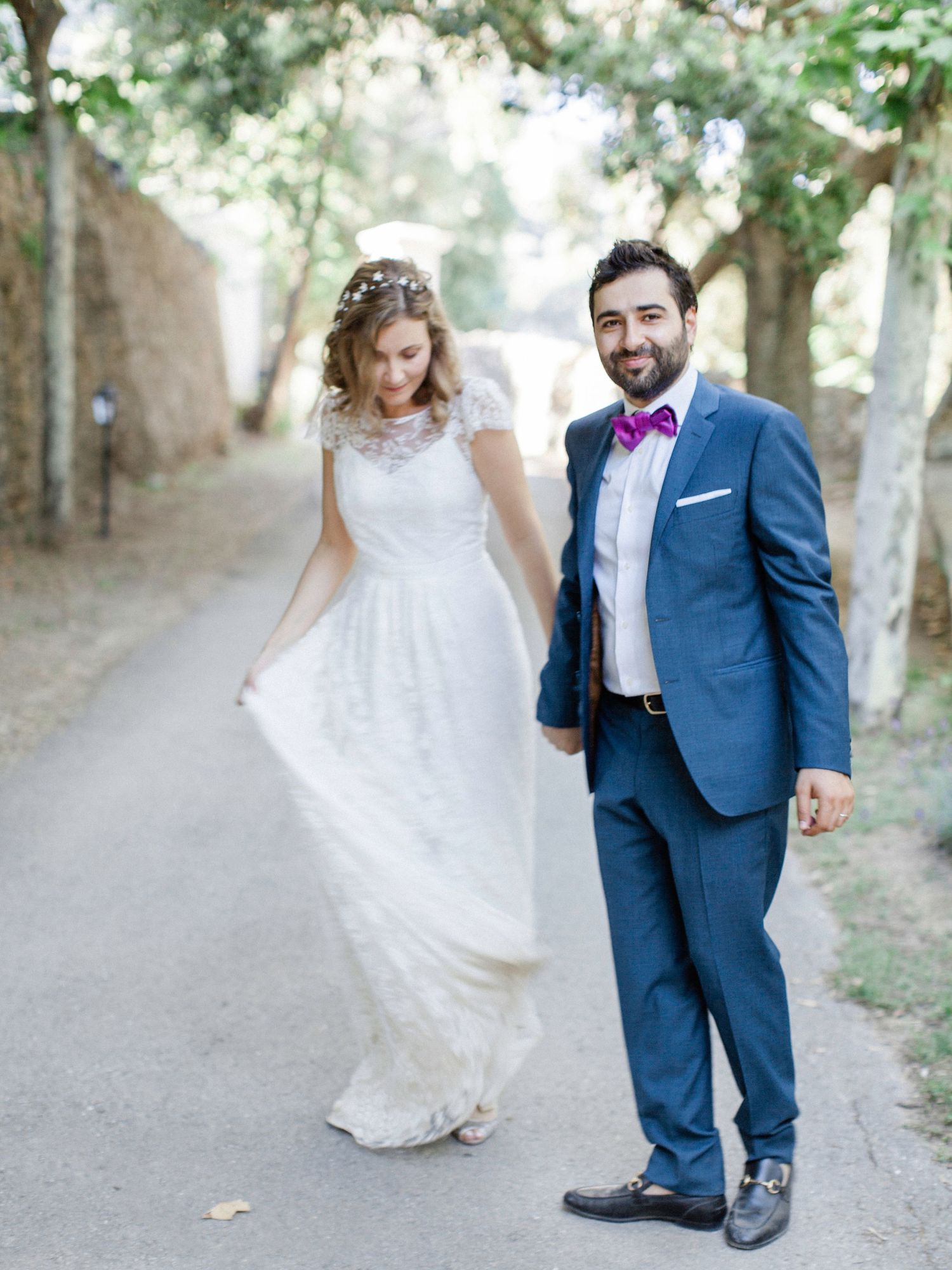 Bride in white lace gown and groom in blue suit with purple bow tie walk hand in hand on a tree-lined path.