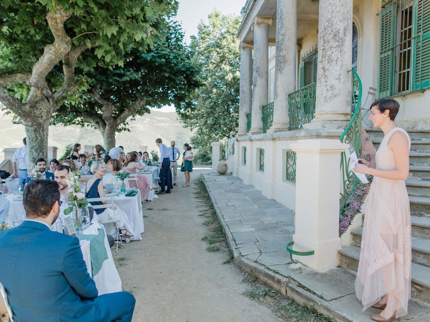 Elegant outdoor wedding reception at a historic villa with guests dining under trees on a sunny day.