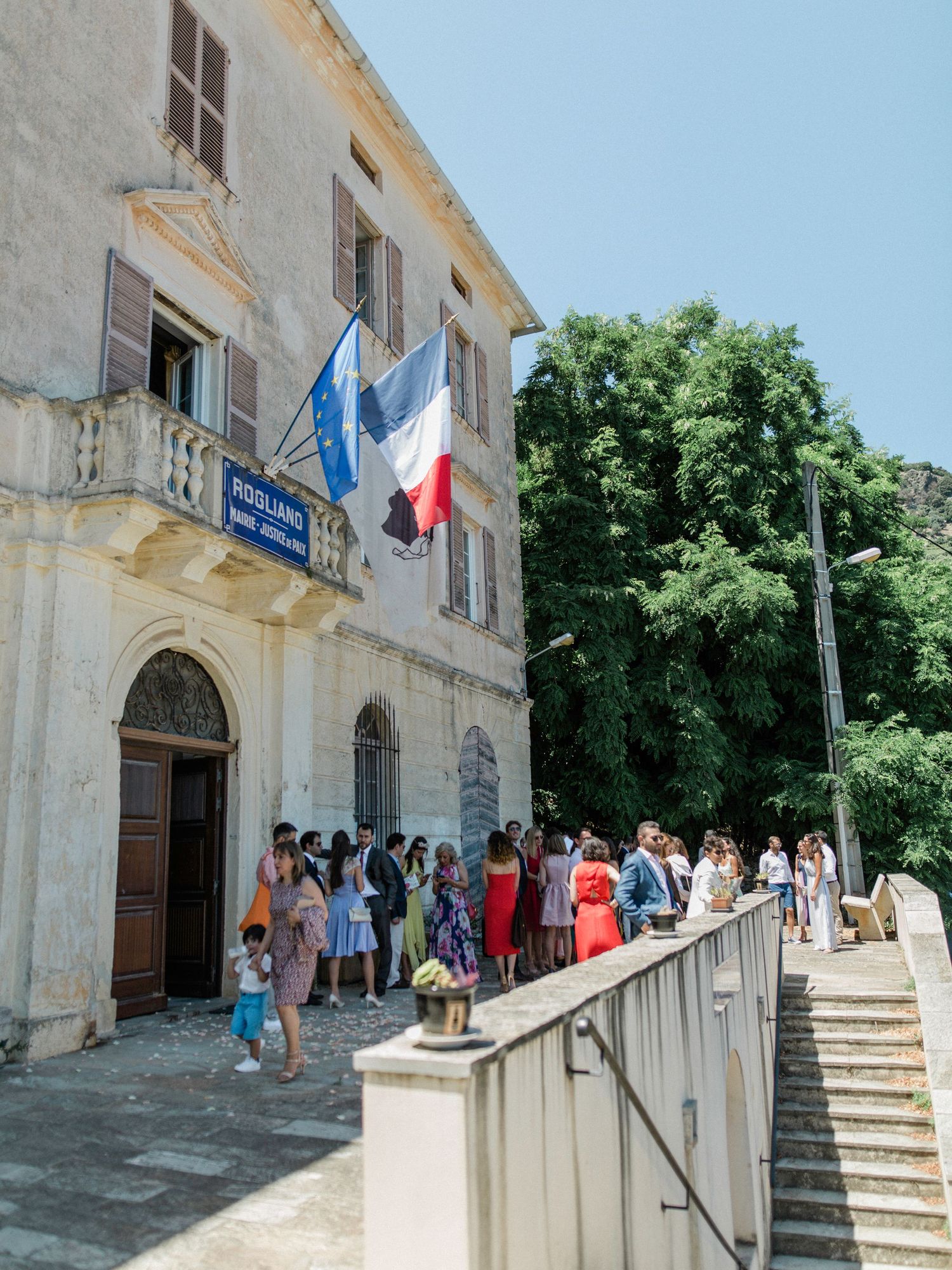 Guests gather outside a historic French building with flags for a sunny outdoor wedding ceremony.