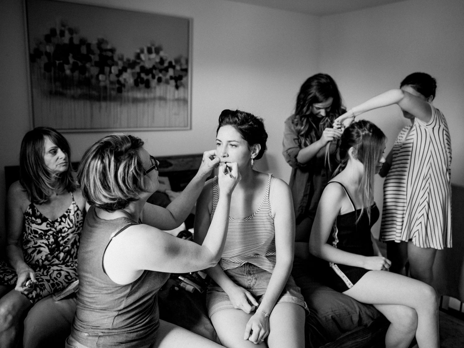 Black and white photo of a bride getting hair and makeup done by bridesmaids in a hotel room before her wedding.