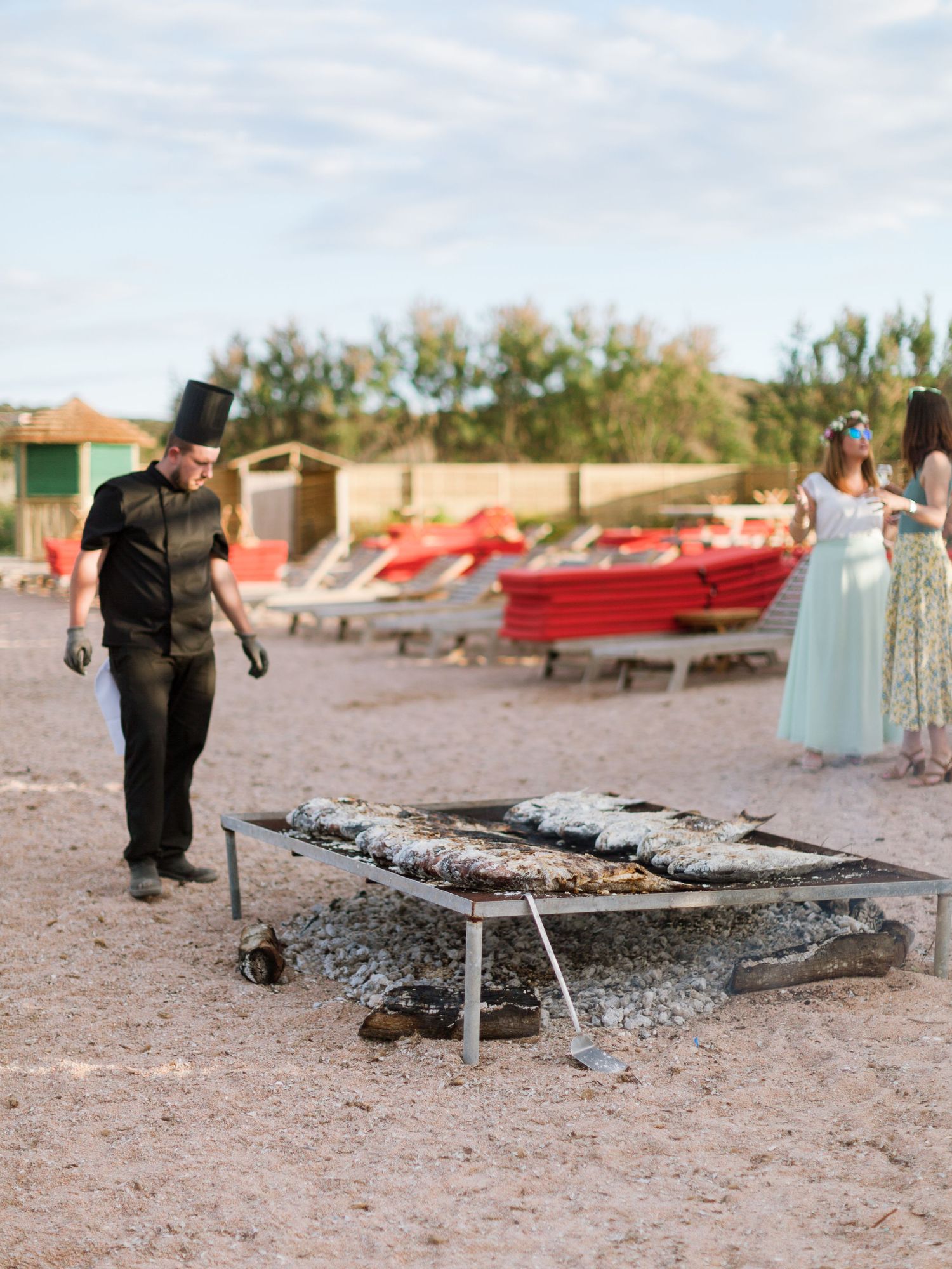Un chef en uniforme noir fait griller du poisson frais sur un barbecue en plein air, en bord de plage, avec des bateaux rouges