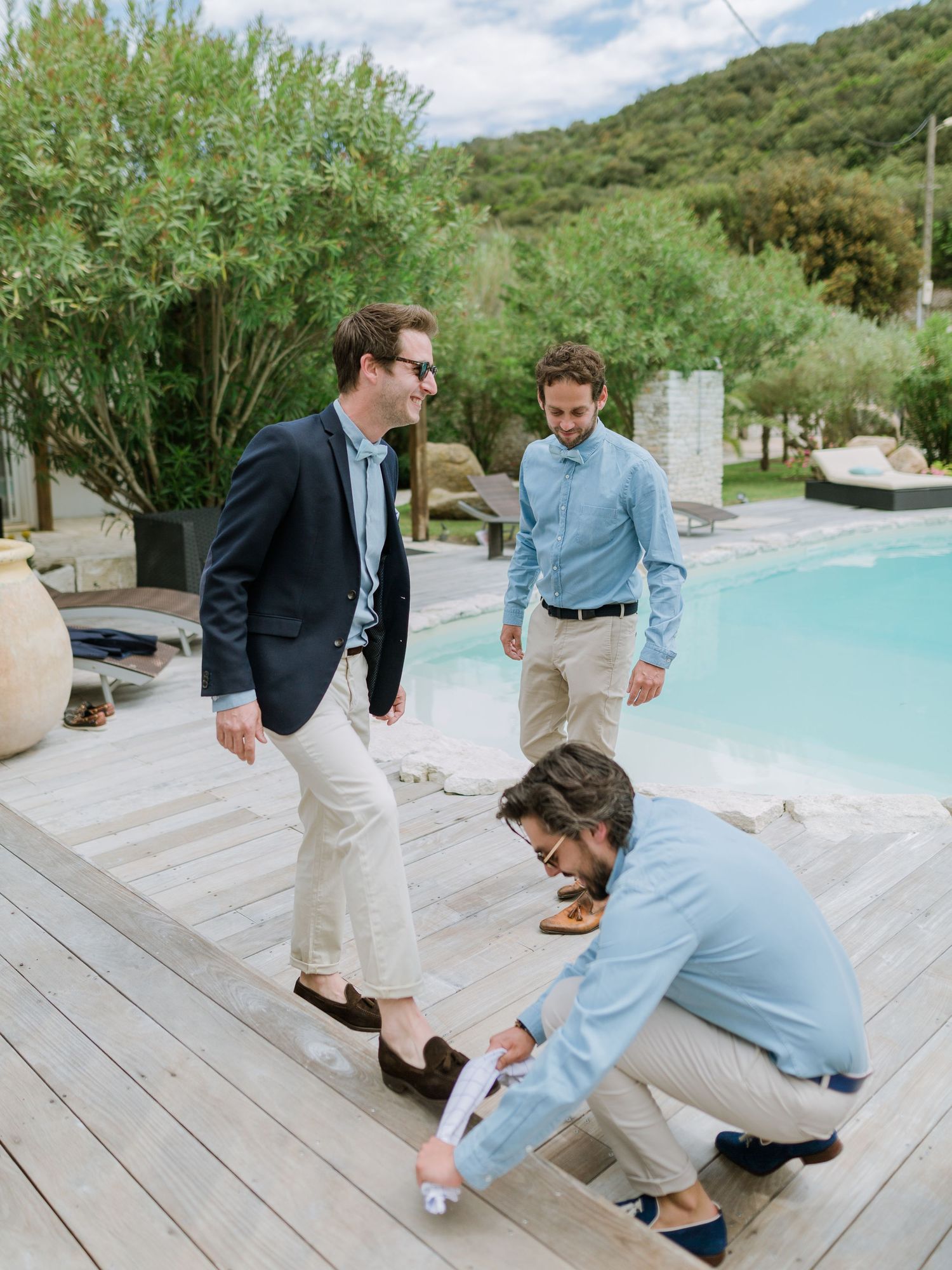 Trois hommes élégants en costumes clairs discutent sur une terrasse de piscine en bois entourée d'une végétation luxuriante.