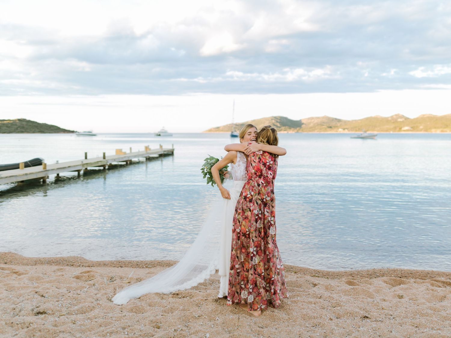Une mariée en robe blanche enlace une femme en robe à fleurs sur une plage de sable fin, avec une jetée et l'eau bleue en arrière-plan.