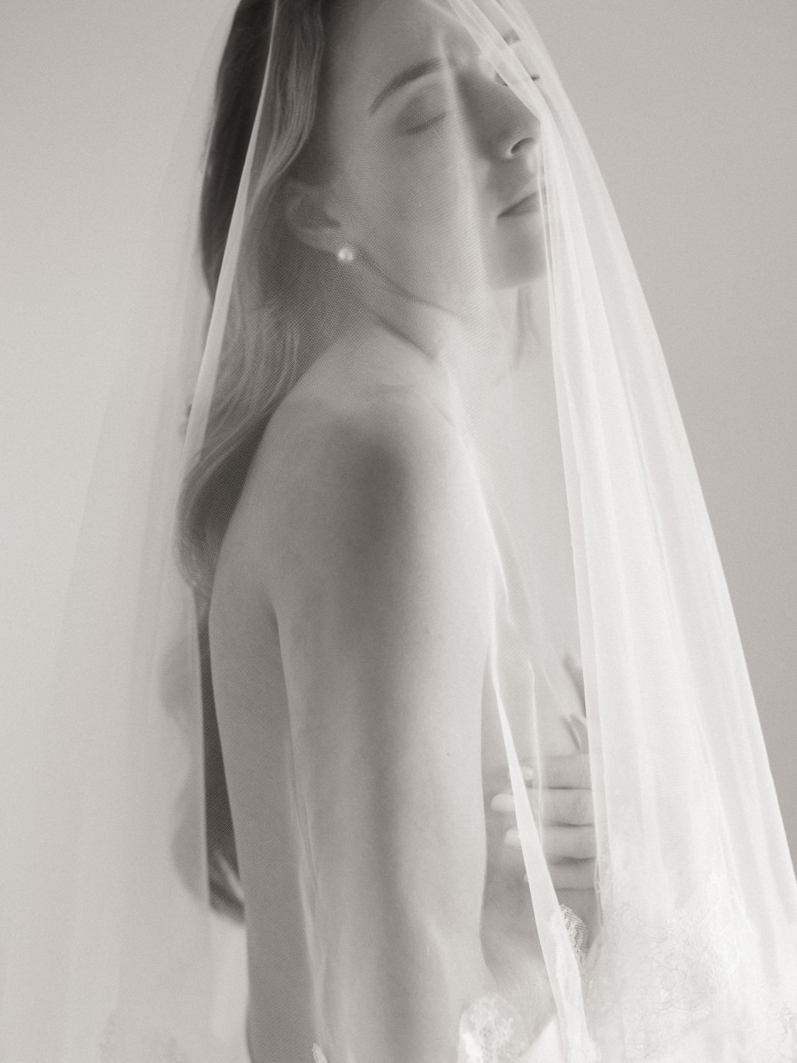 Black and white bridal portrait of a woman in a wedding veil with eyes closed, wearing a strapless gown and pearl earring.