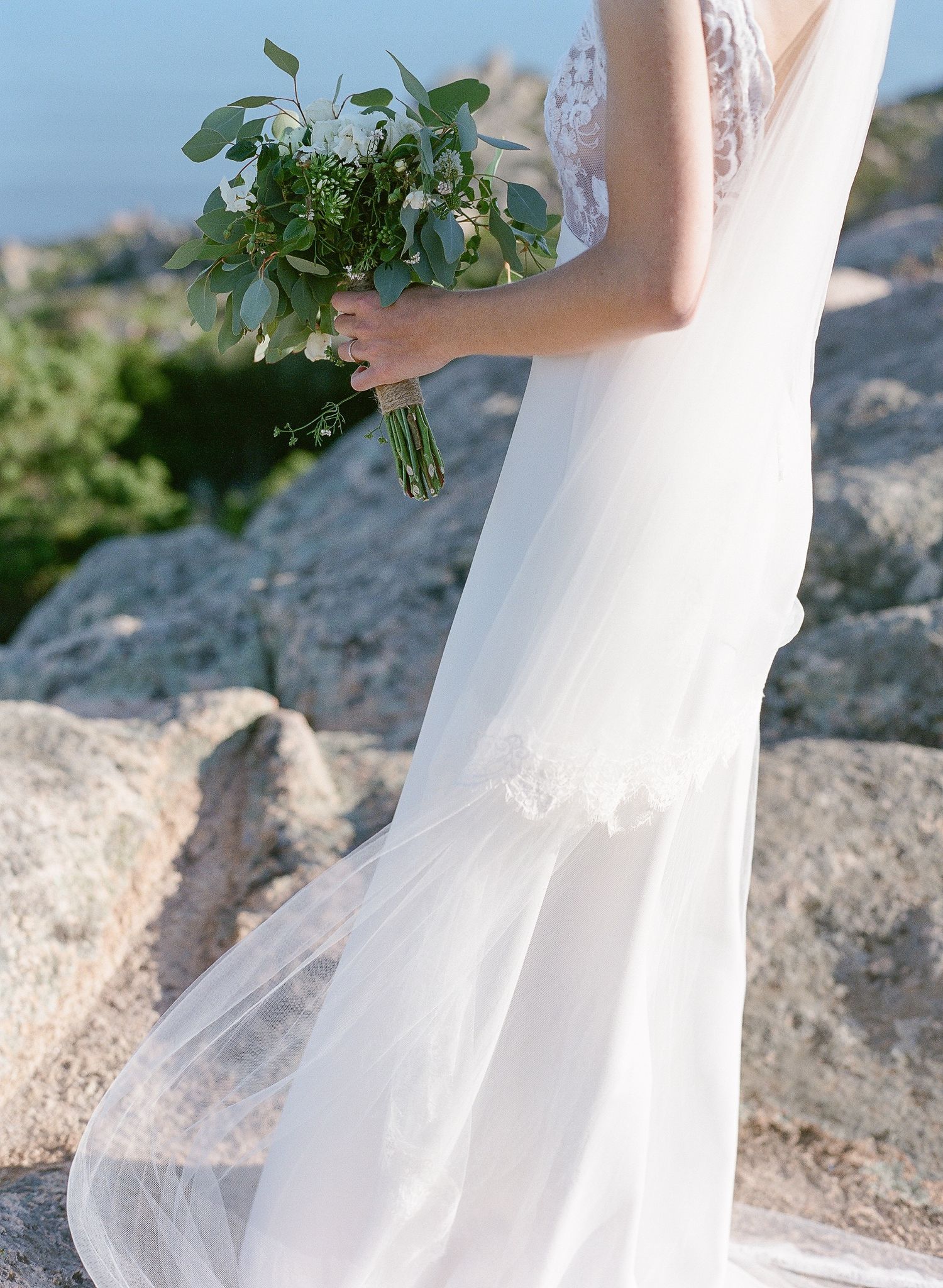 Une mariée en robe de dentelle fluide tient un bouquet d'eucalyptus vert dans un décor côtier rocheux. A la trinité de Bonifacio.