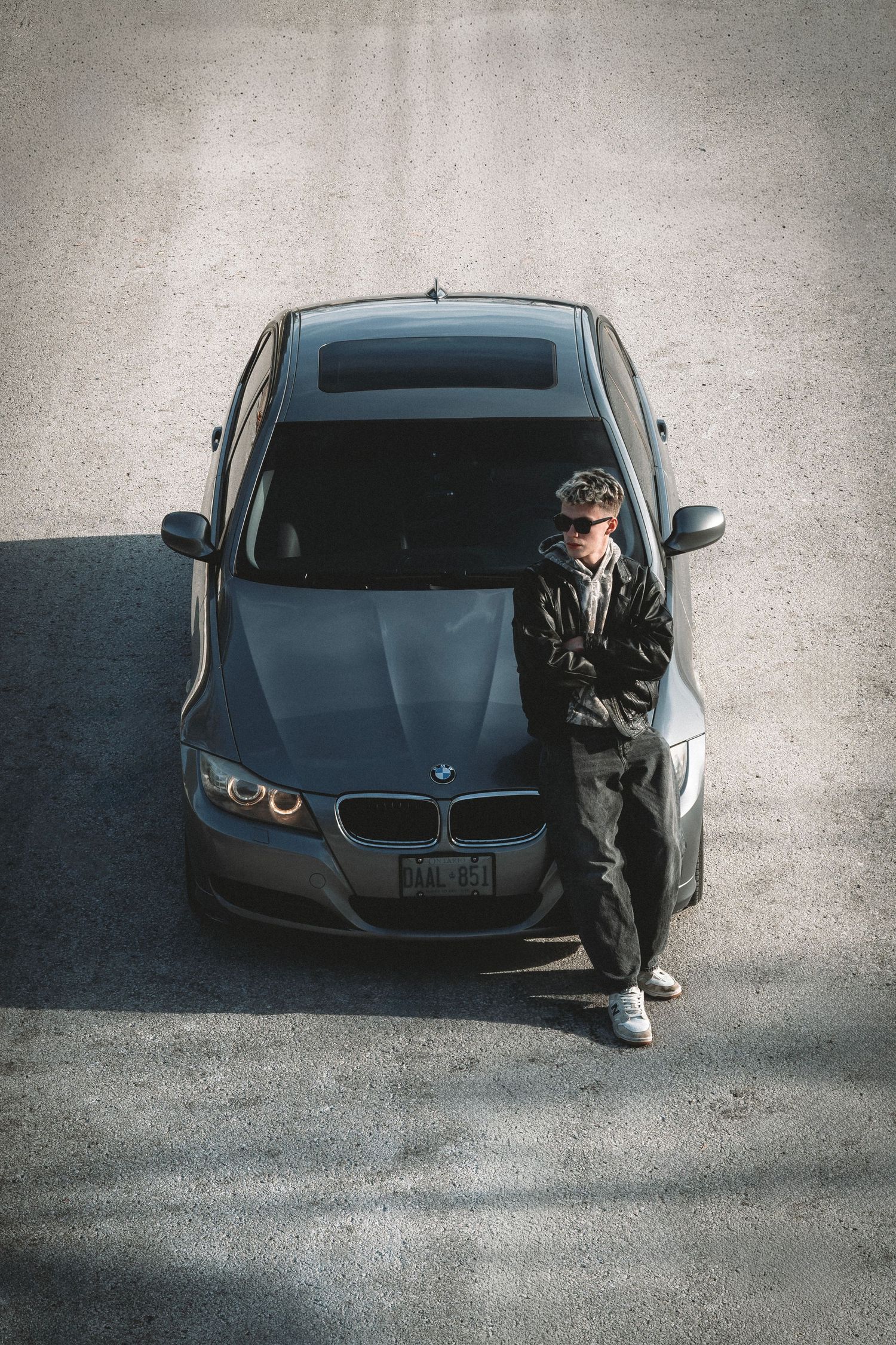 Photographer leaning against a gray BMW 3 Series, shot from above in a parking lot.