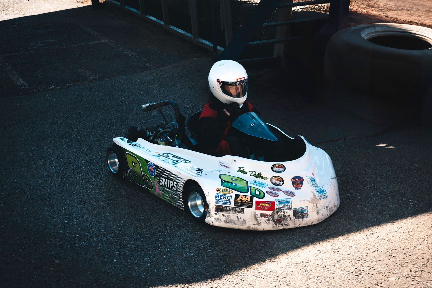 A driver wearing a helmet sitting in a small white go-kart covered in stickers on an outdoor track.