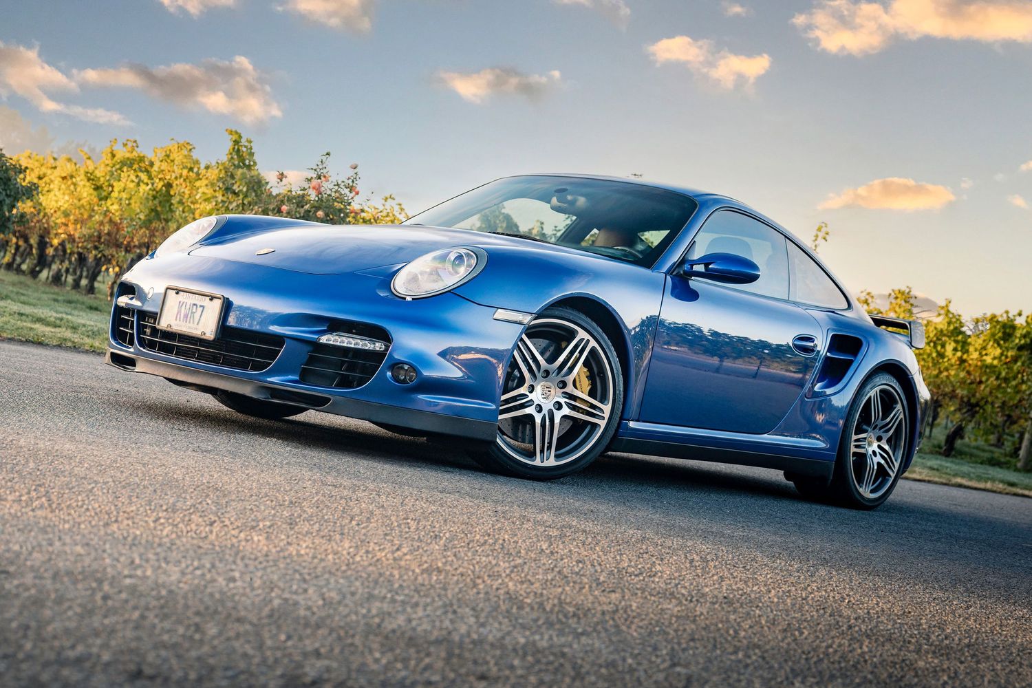A blue Porsche 911 Turbo on a road with autumn foliage and a dramatic cloudy sky.