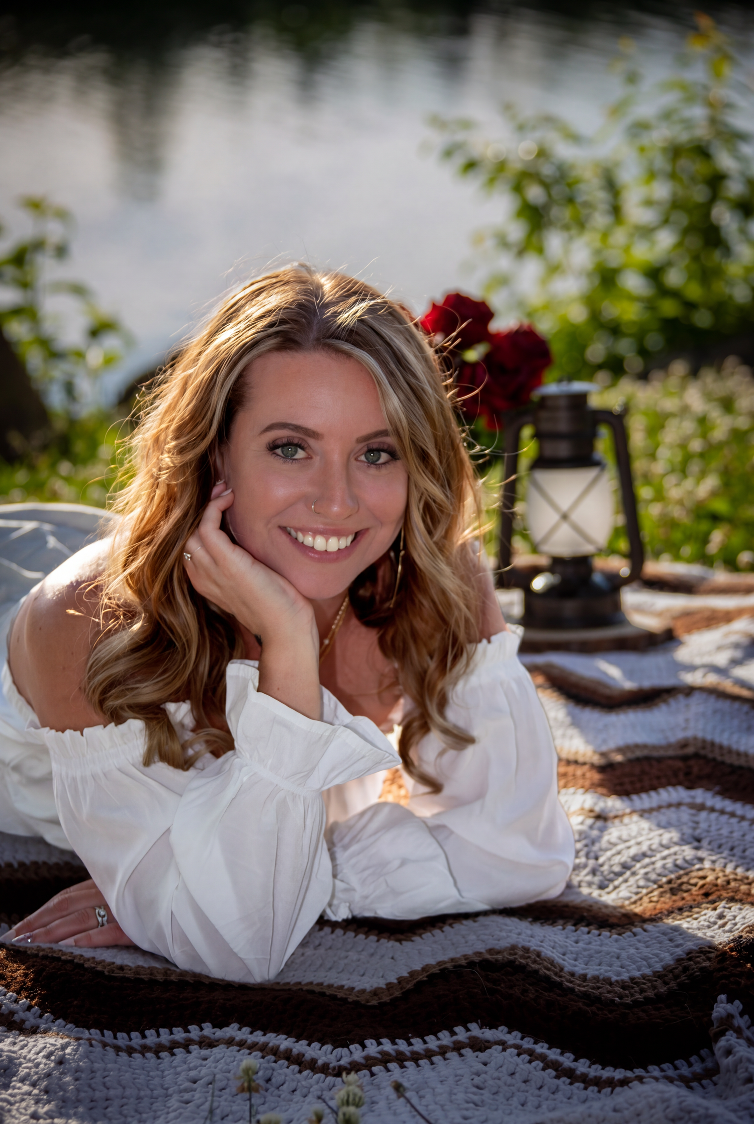 Smiling woman in white off-shoulder dress lying on crochet blanket by a serene lake with red roses.