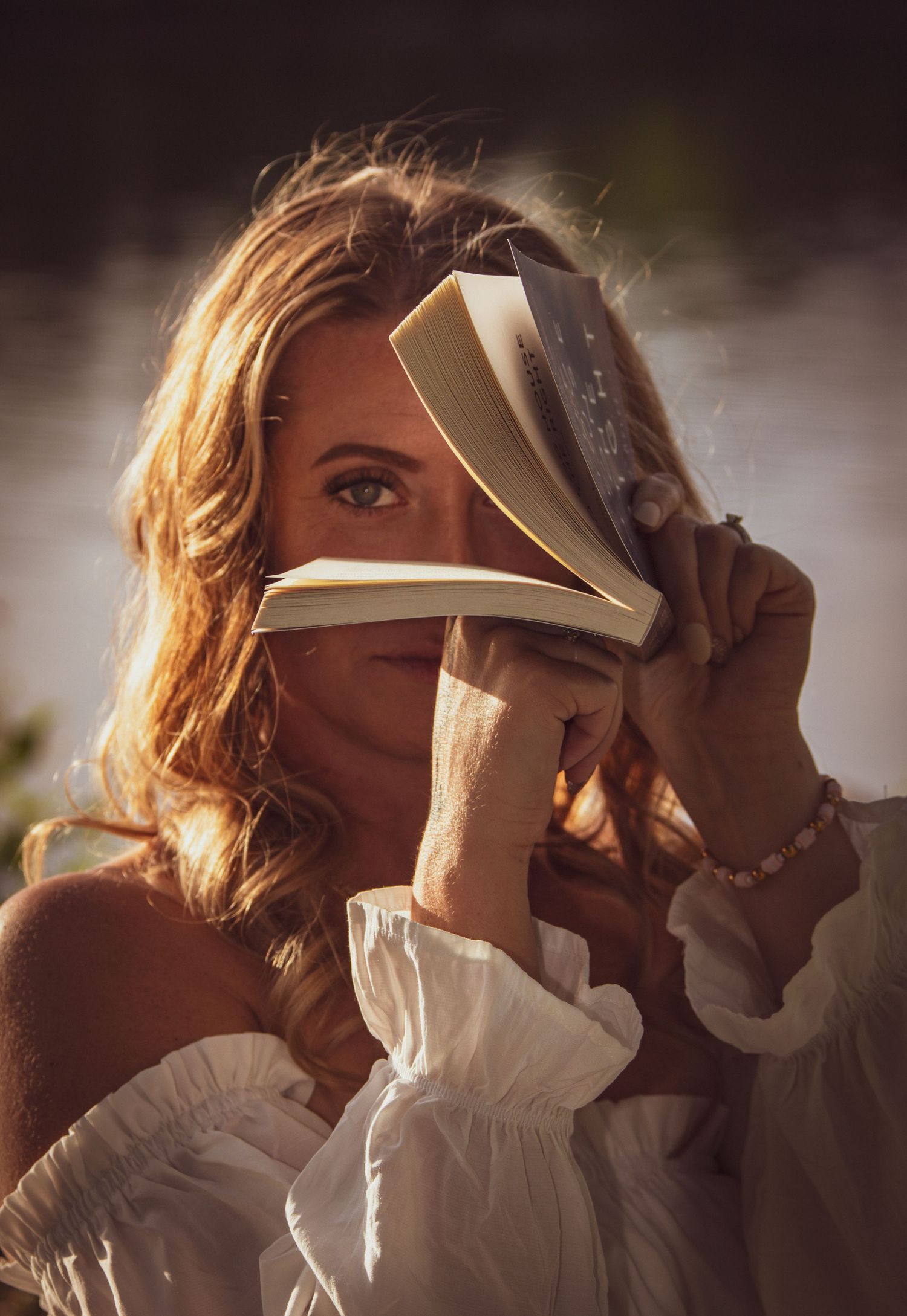 Woman in white off-shoulder top holding open book in front of her face in golden sunlight.