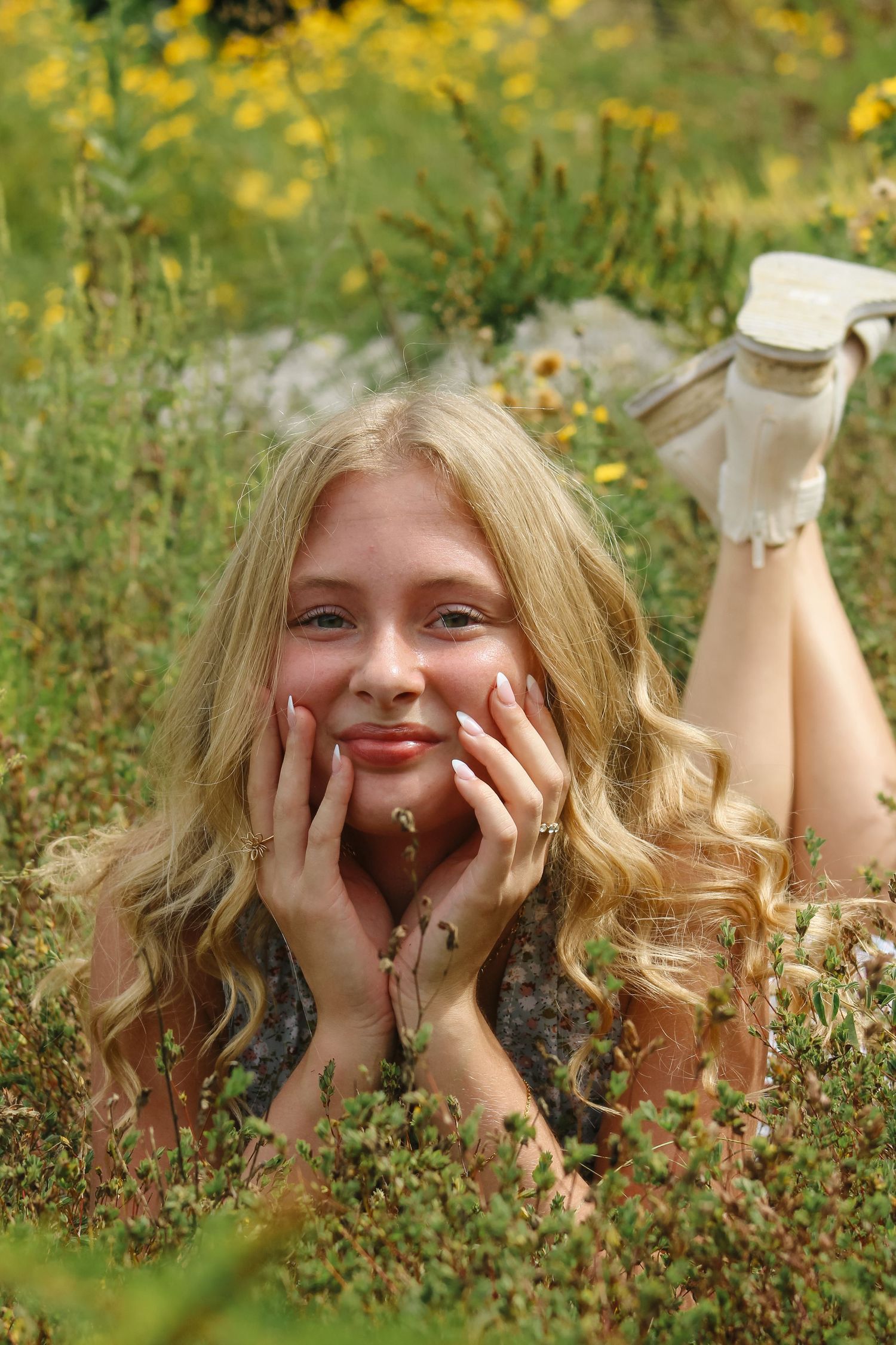Young woman lying in grassy field with wildflowers, hands on cheeks, legs raised behind her during outdoor portrait session