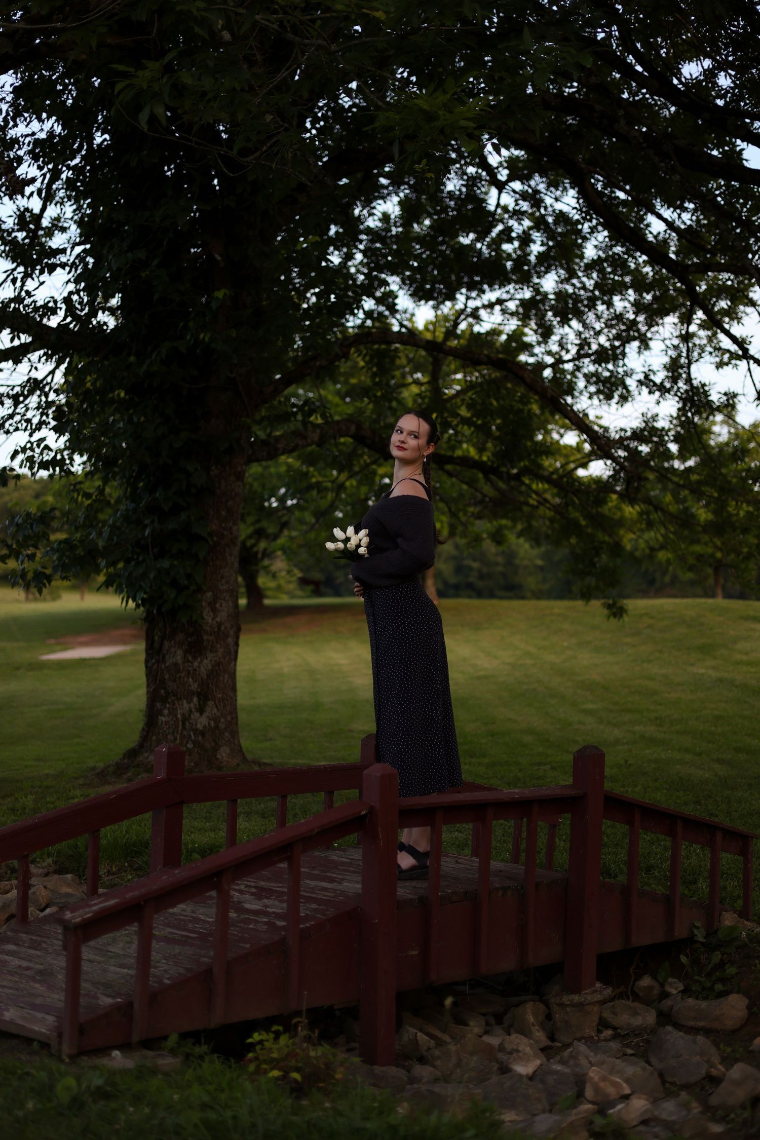 Young woman standing on small bridge under tree shade with large tree in background during outdoor portrait session