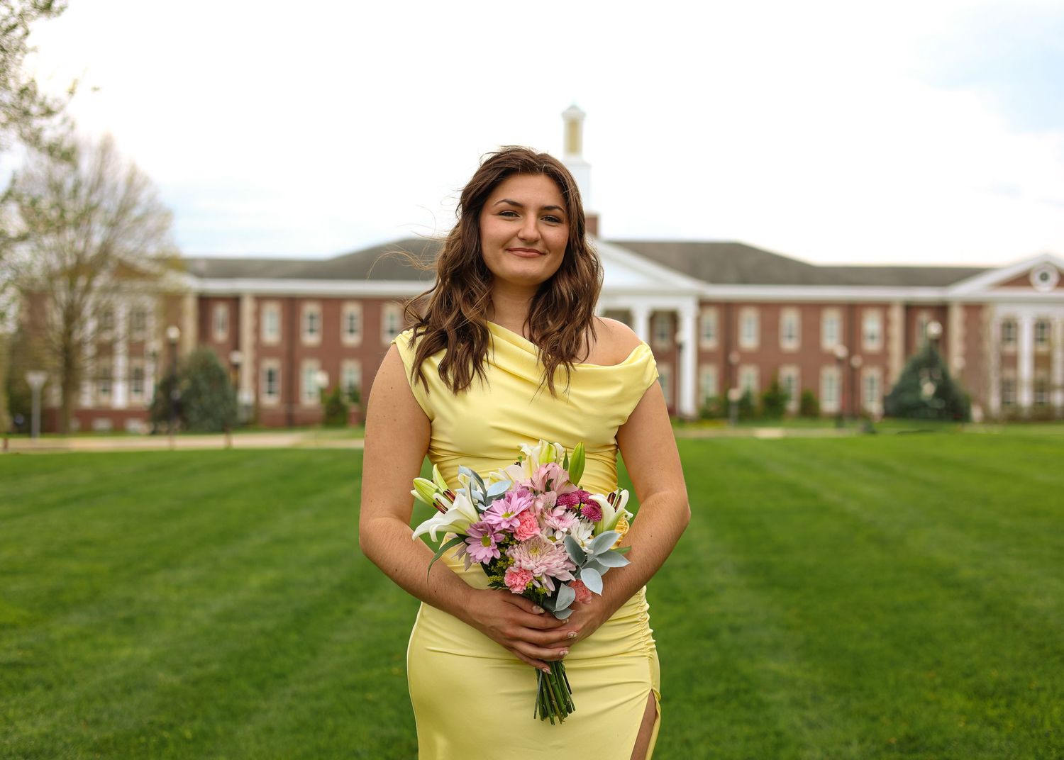 College graduate standing on Tennessee Tech lawn holding flowers with Glory Bell Hall nursing building in the background