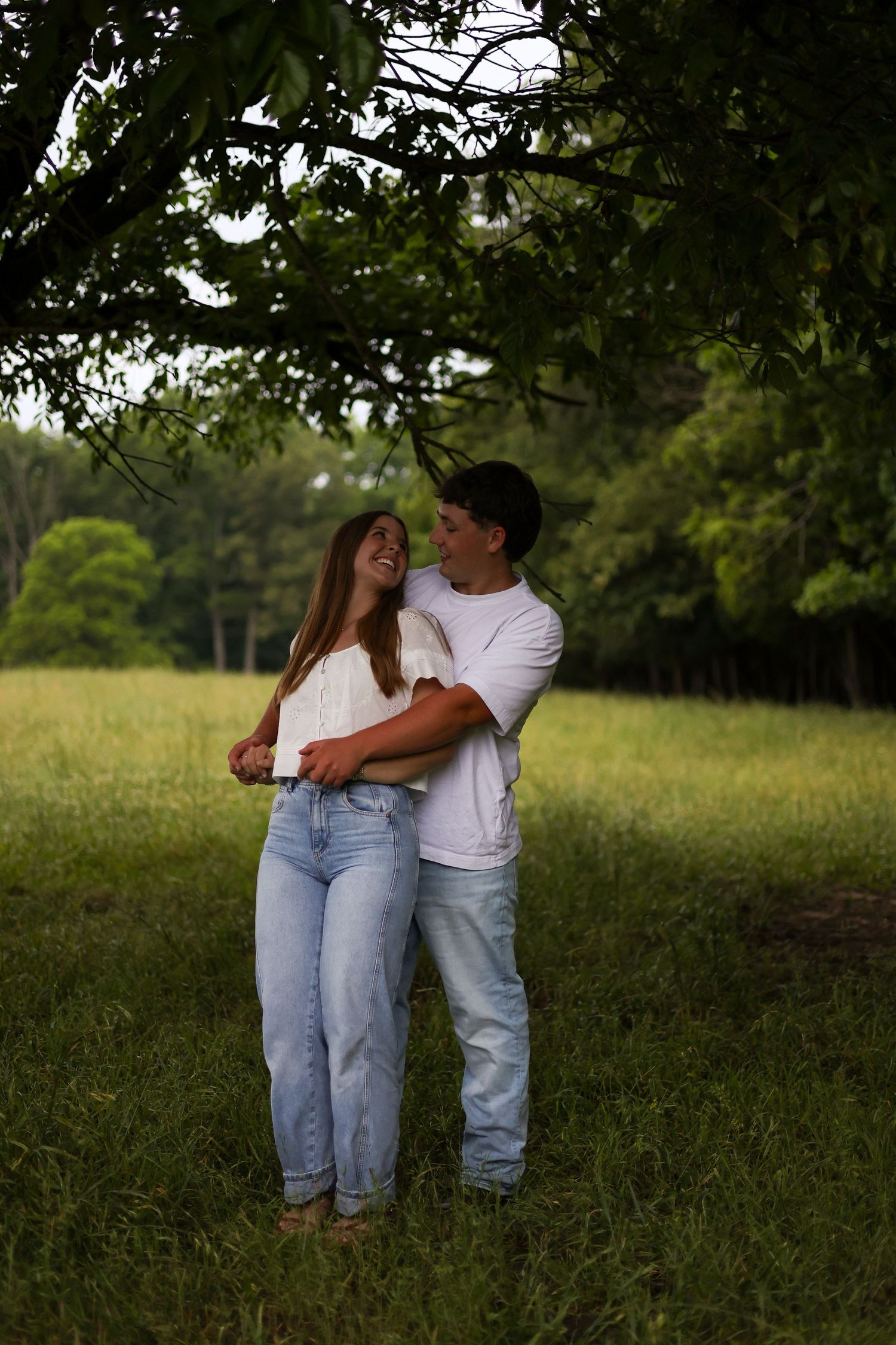 Couple in white shirts and jeans standing under a tree in a green field, looking at each other during outdoor portrait session