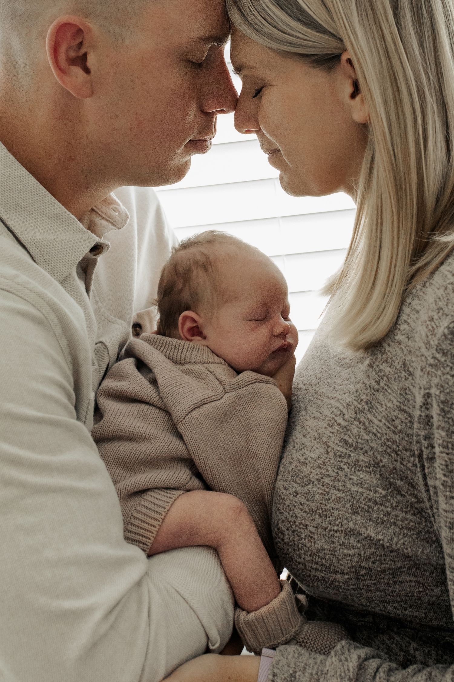 New parents tenderly touch foreheads while cradling their sleeping newborn baby dressed in a beige knit outfit.