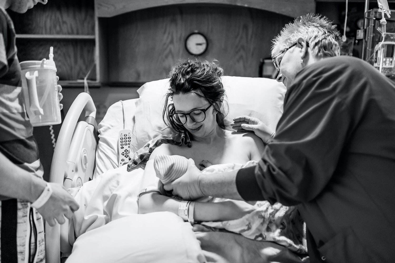 New mother holds newborn baby in hospital bed surrounded by family, emotional black and white birth photo.