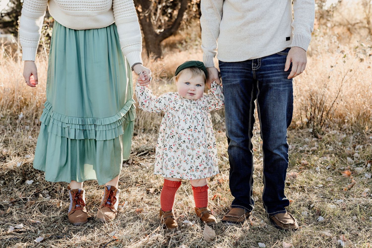 Smiling toddler in floral dress and green headband holding parents hands in an autumn field.