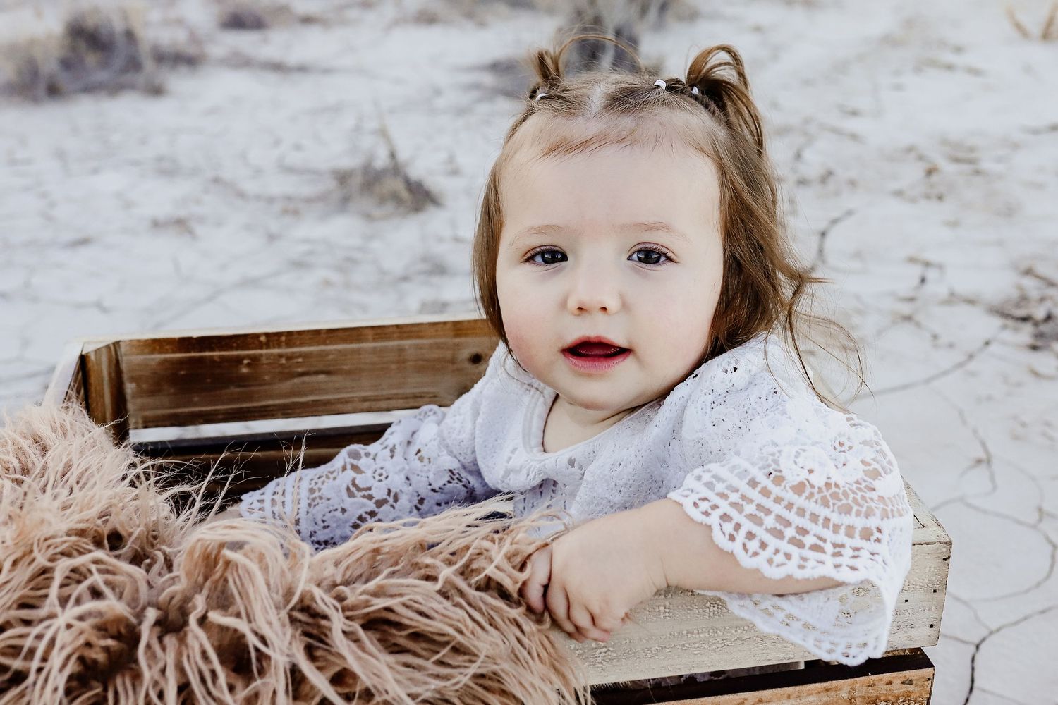 Adorable baby girl with big eyes sitting inside a rustic wooden crate with burlap on sandy ground.