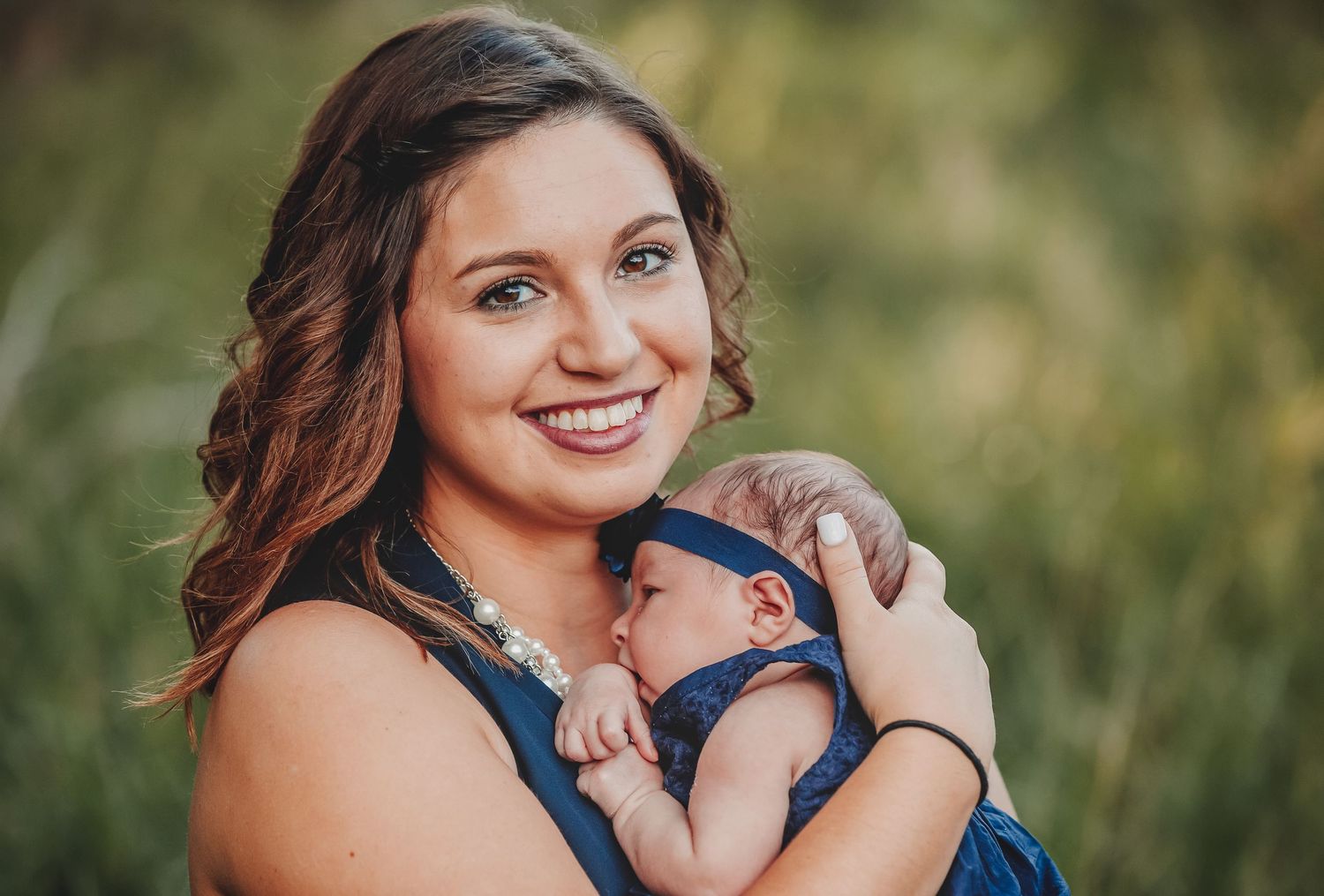 Smiling young mother holding her sleeping newborn baby outdoors in a lush green setting.