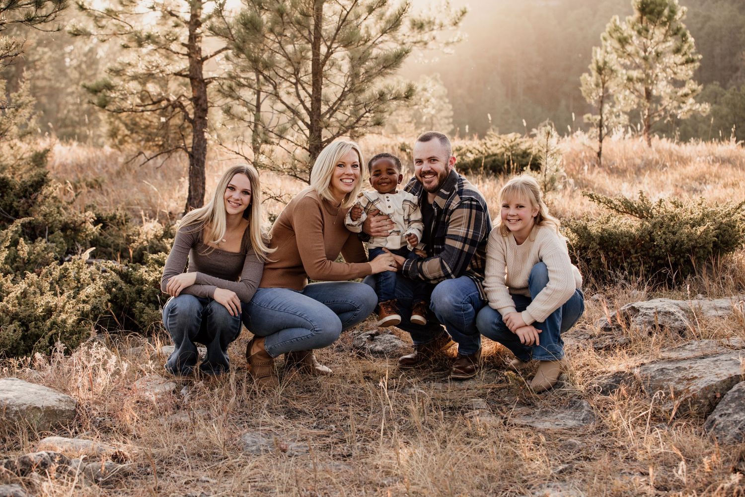 Family of five smiling outdoors in a sunlit forest setting during autumn, posing together on rocky ground.