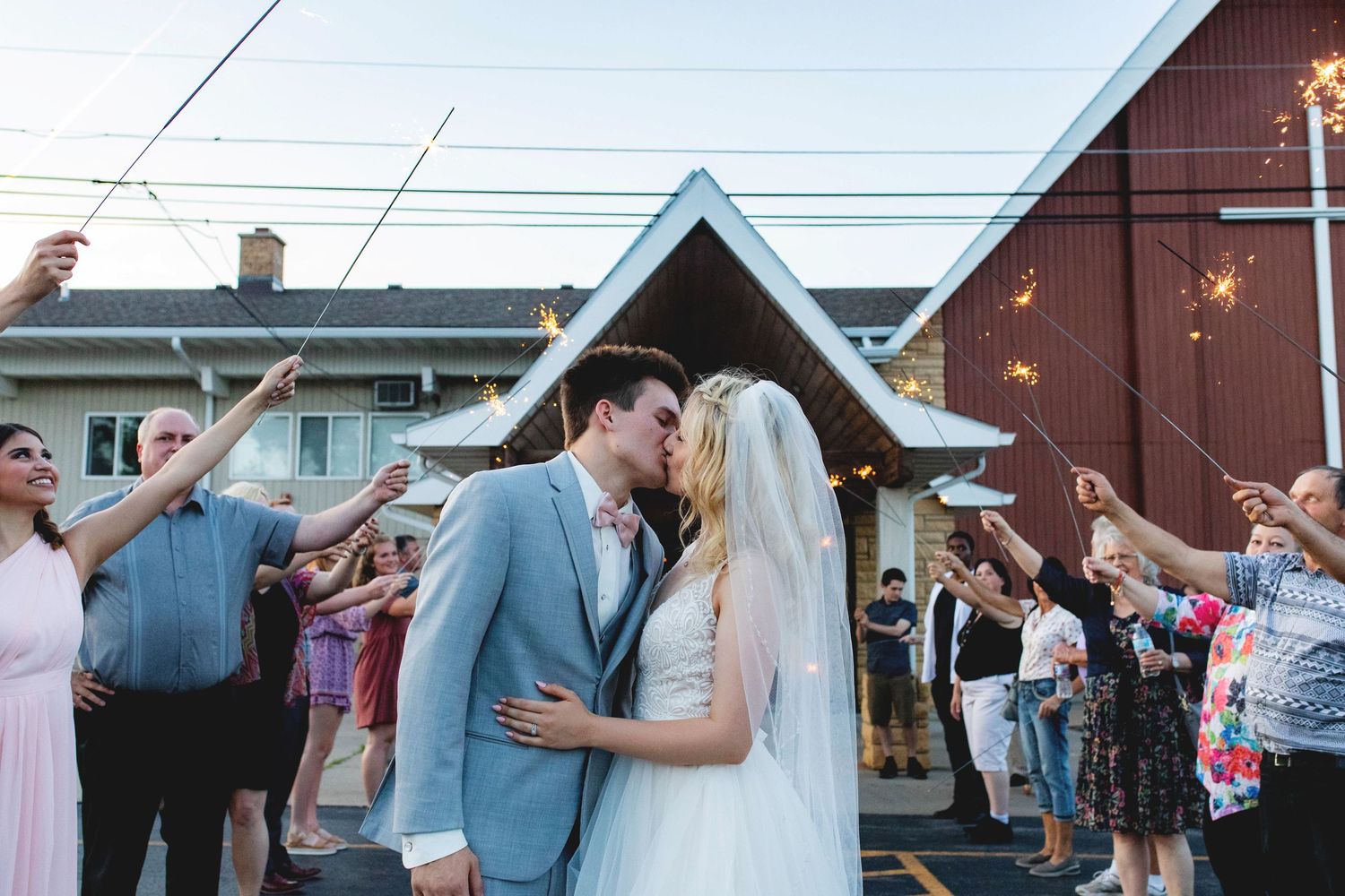 Newlywed couple kissing during sparkler send-off at outdoor wedding reception with guests celebrating around them.