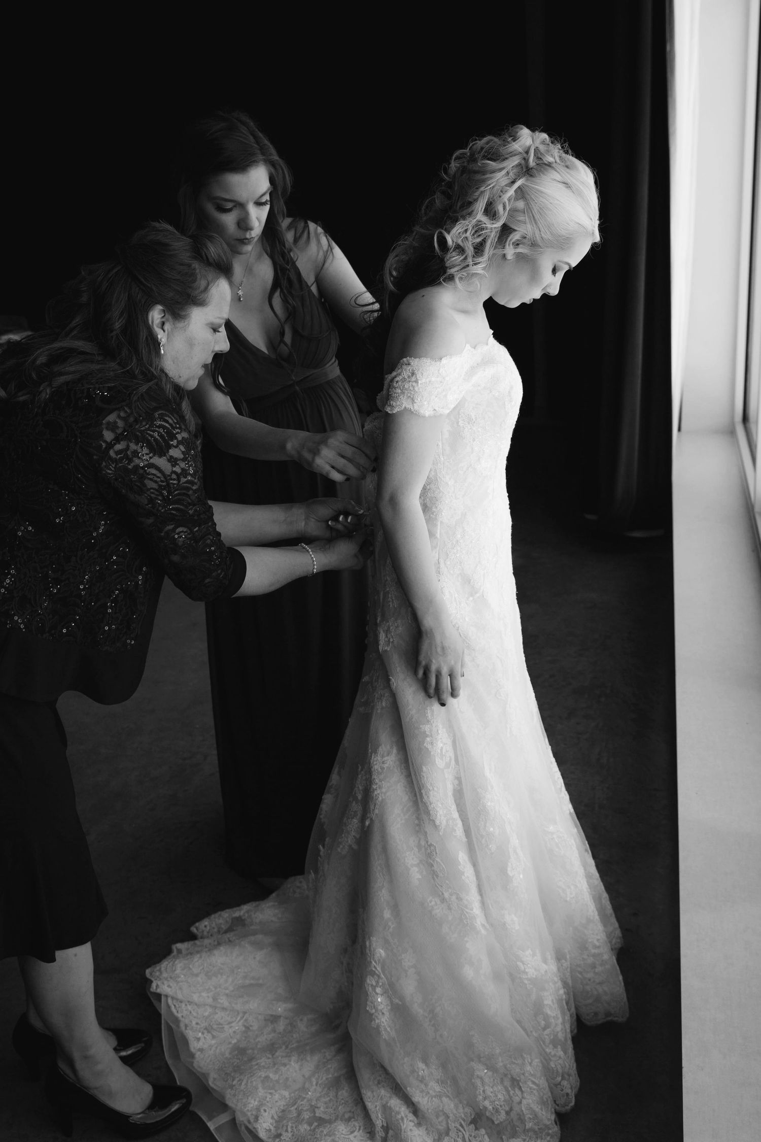 Two bridesmaids help a bride into her lace wedding gown in this elegant black and white bridal preparation photo.