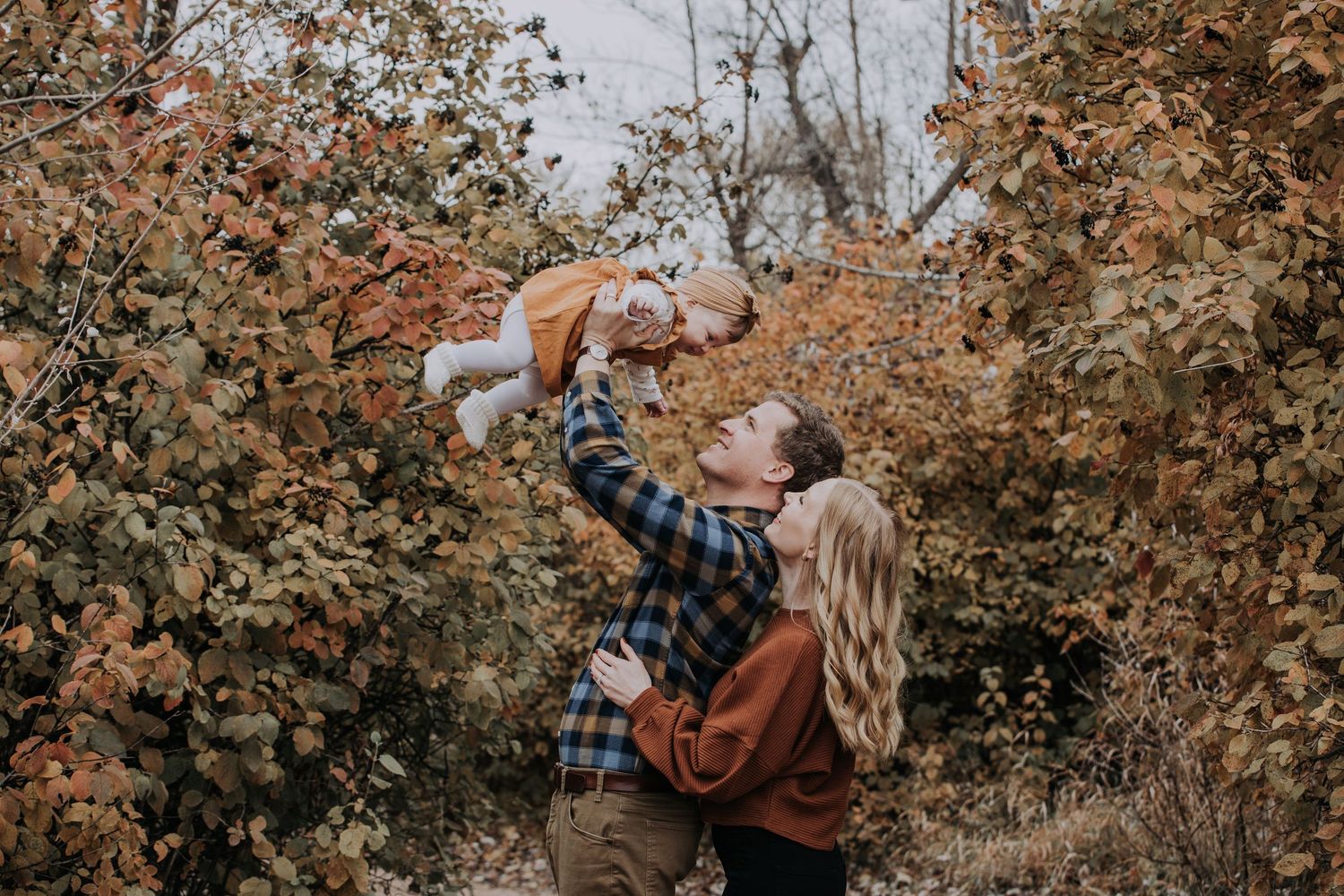 A couple lifts their baby in autumn foliage, sharing a joyful family moment outdoors.