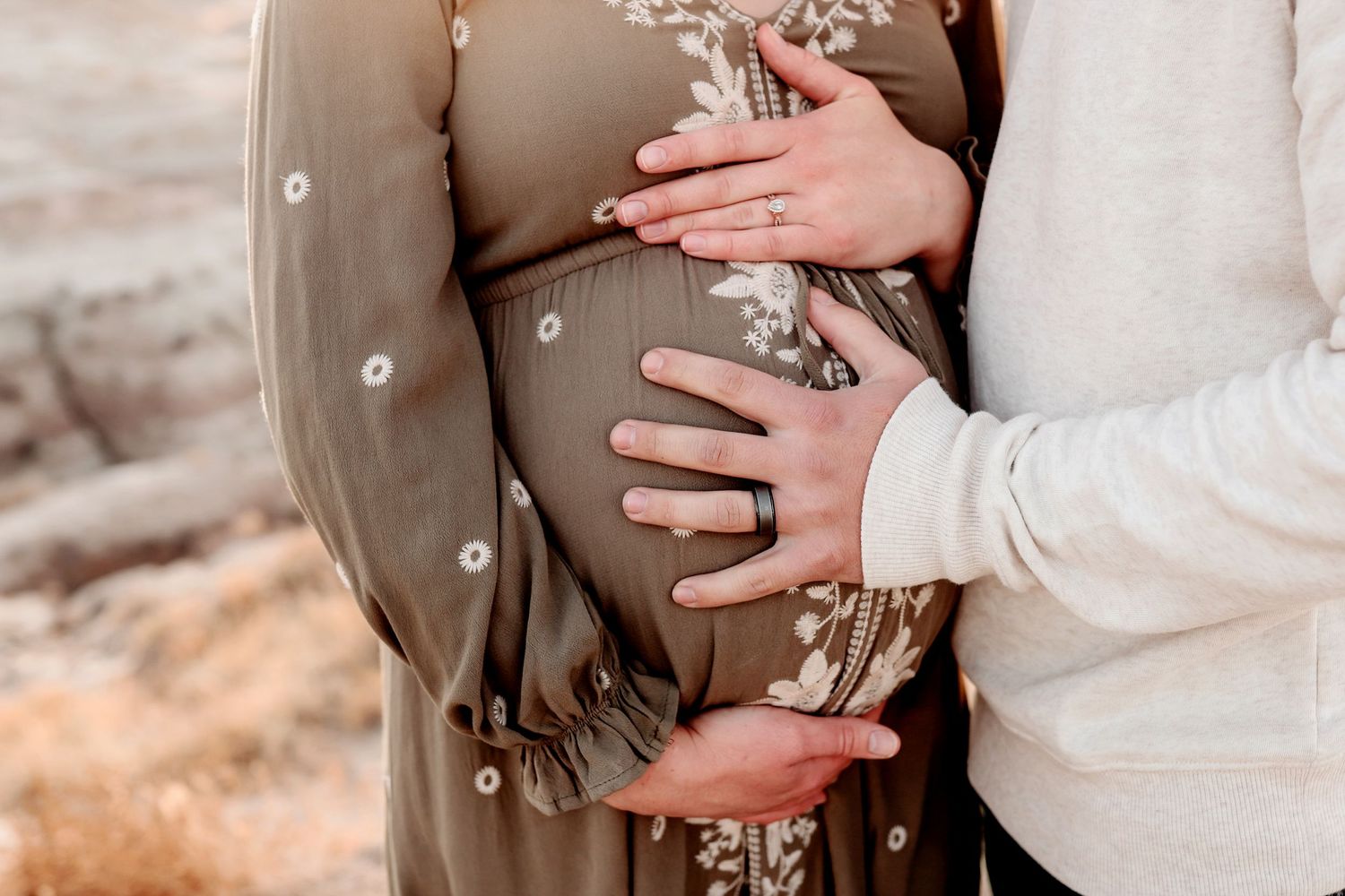 Partner embracing pregnant woman in floral dress from behind, both cradling baby bump outdoors.