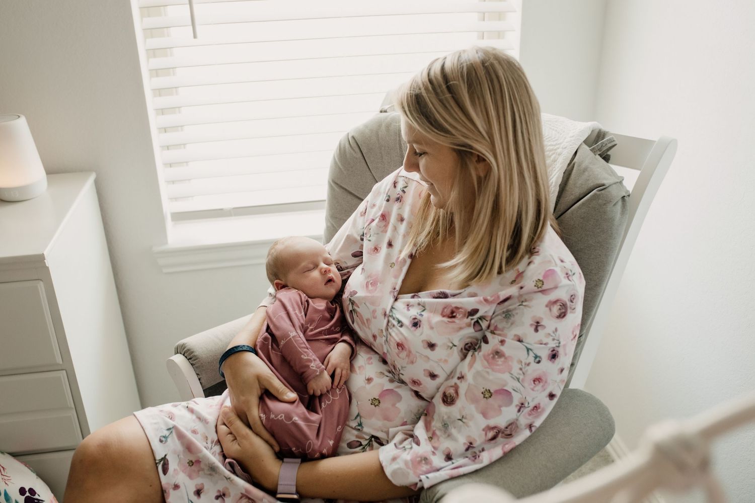Blonde mother in floral robe sitting in nursery glider lovingly gazing at newborn baby.