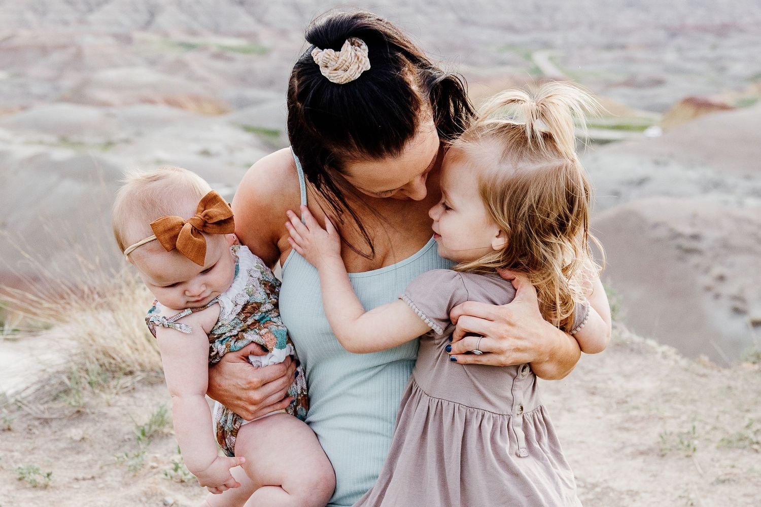 Mother tenderly embracing two young daughters outdoors in a scenic desert landscape during a family photo session.