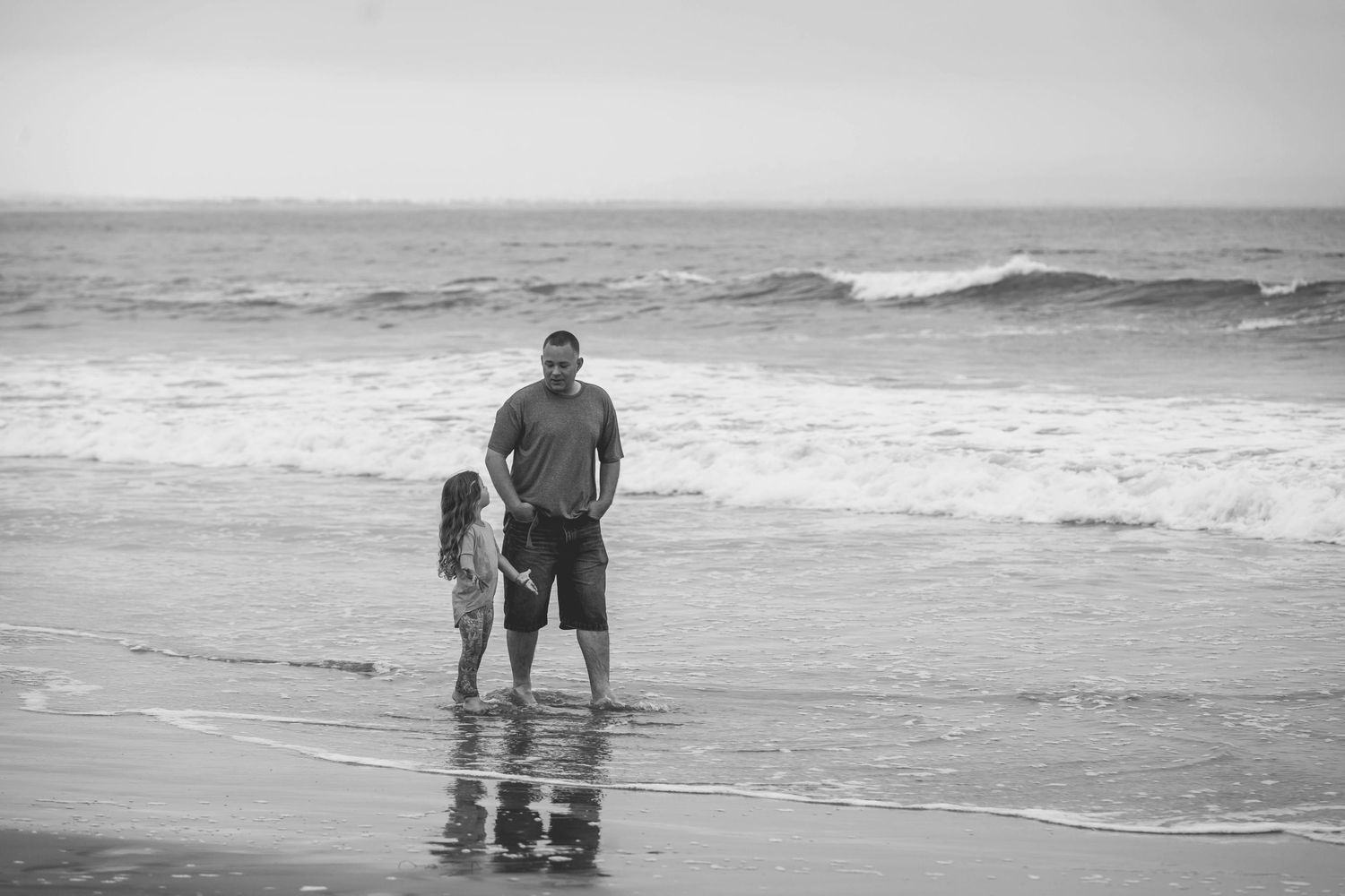 Father and daughter stand together at the waters edge on a beach.