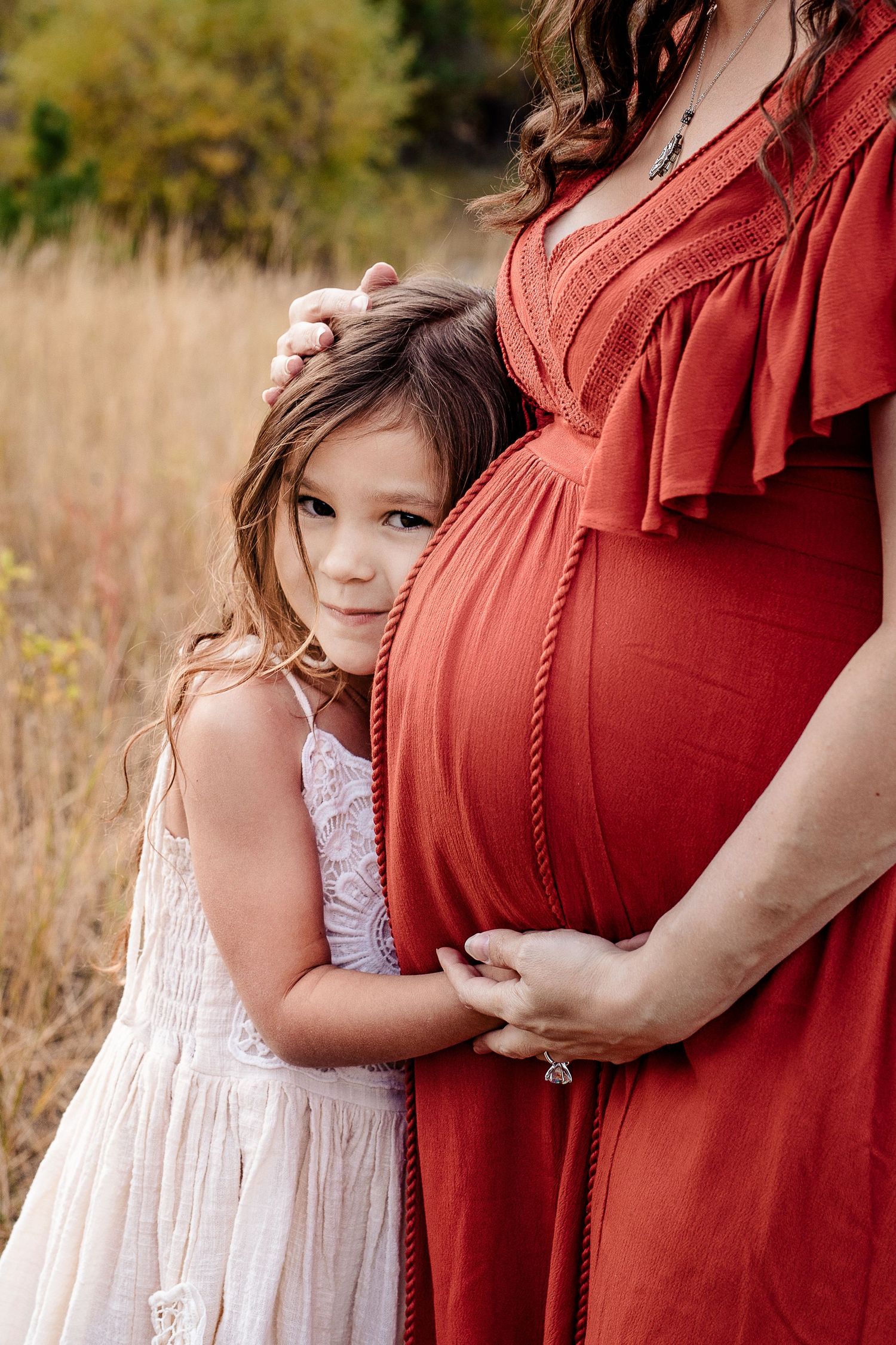 Young girl in white dress lovingly hugs pregnant mothers red-dressed baby bump outdoors.
