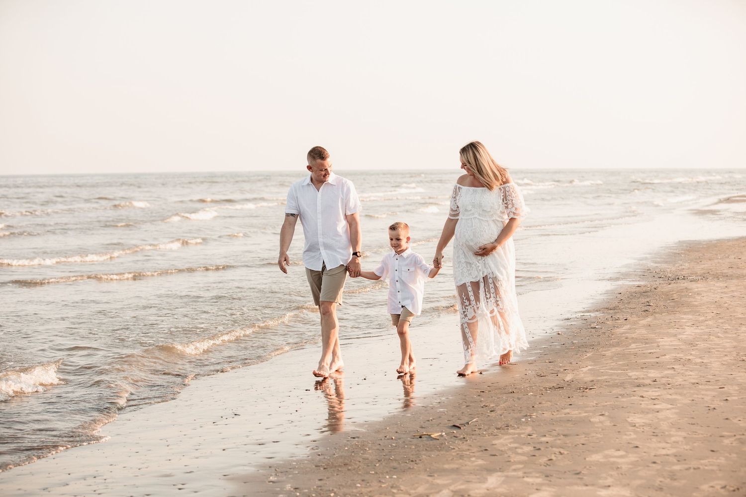 Happy family with pregnant mother, father, and young son walking barefoot along a sunny beach.