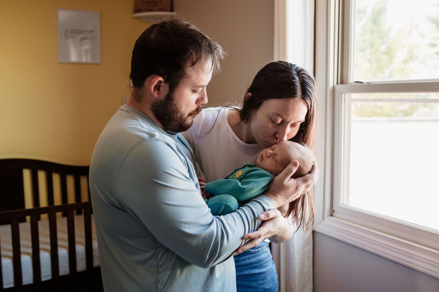 Two parents in a nursery room tenderly hold and gaze at their newborn baby next to a window.