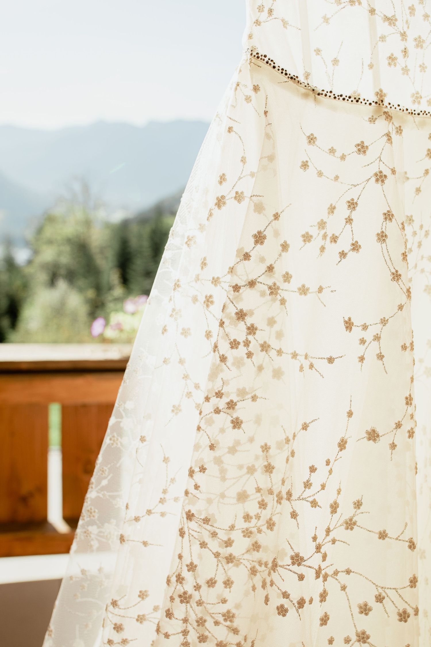Close-up of an ivory lace wedding dress with floral embroidery, hanging on a balcony with mountain views.