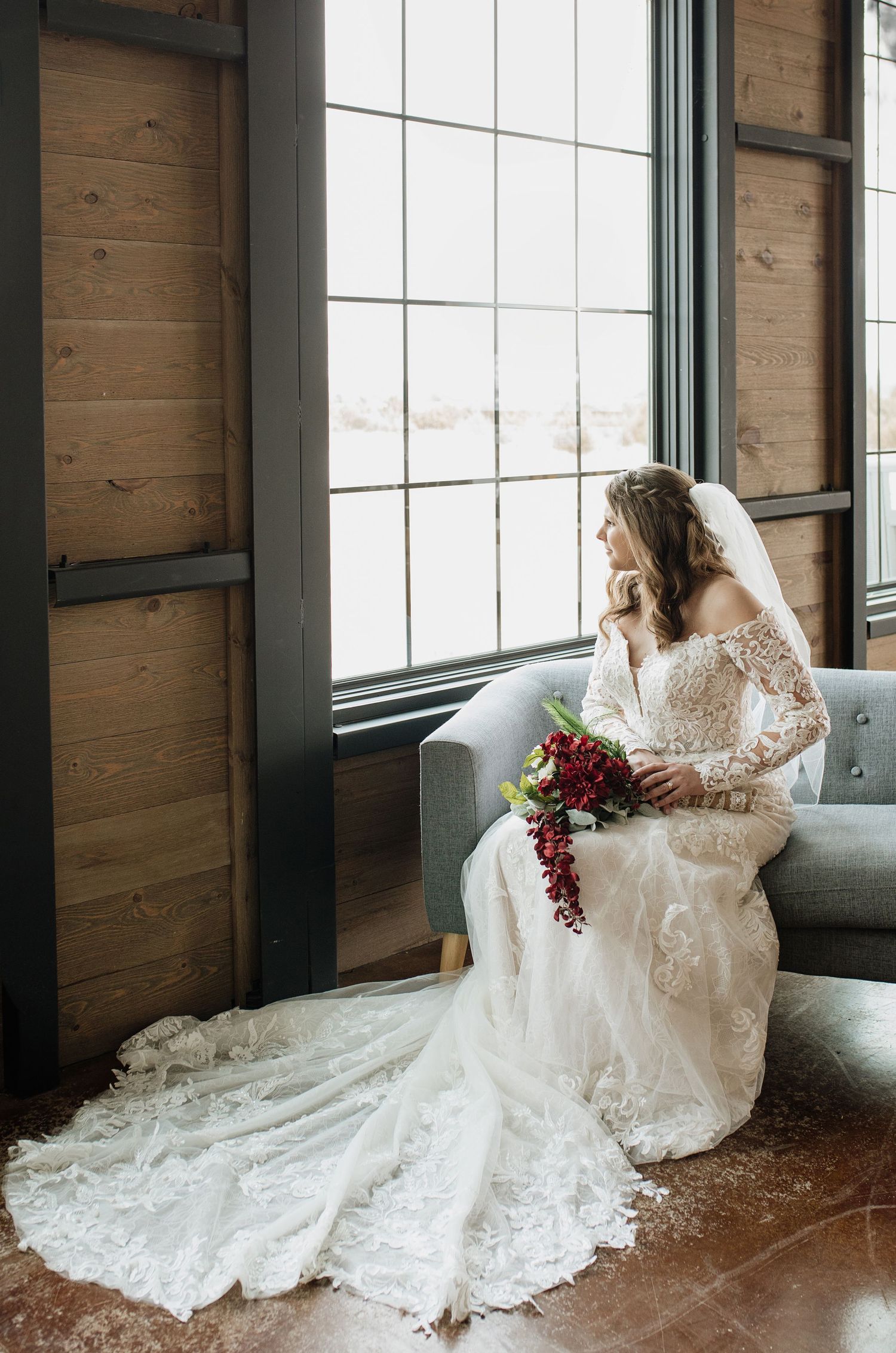 Bride in lace off-shoulder gown with veil holds red bouquet, seated by large window in rustic venue.