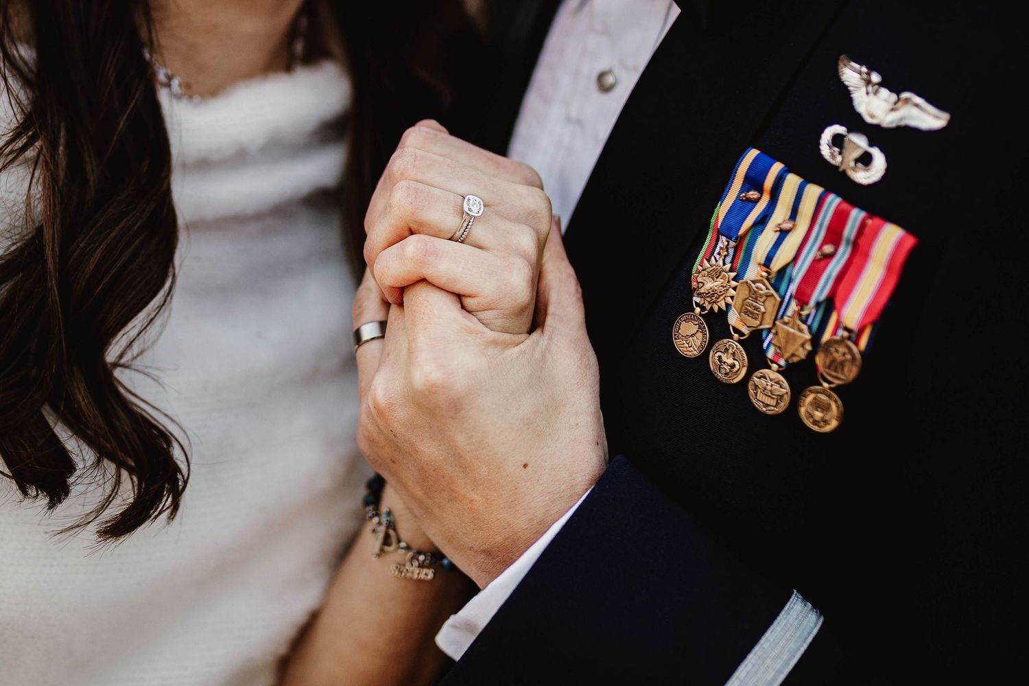 Close-up of a military groom and bride holding hands, showcasing wedding rings and colorful service medals on his uniform.